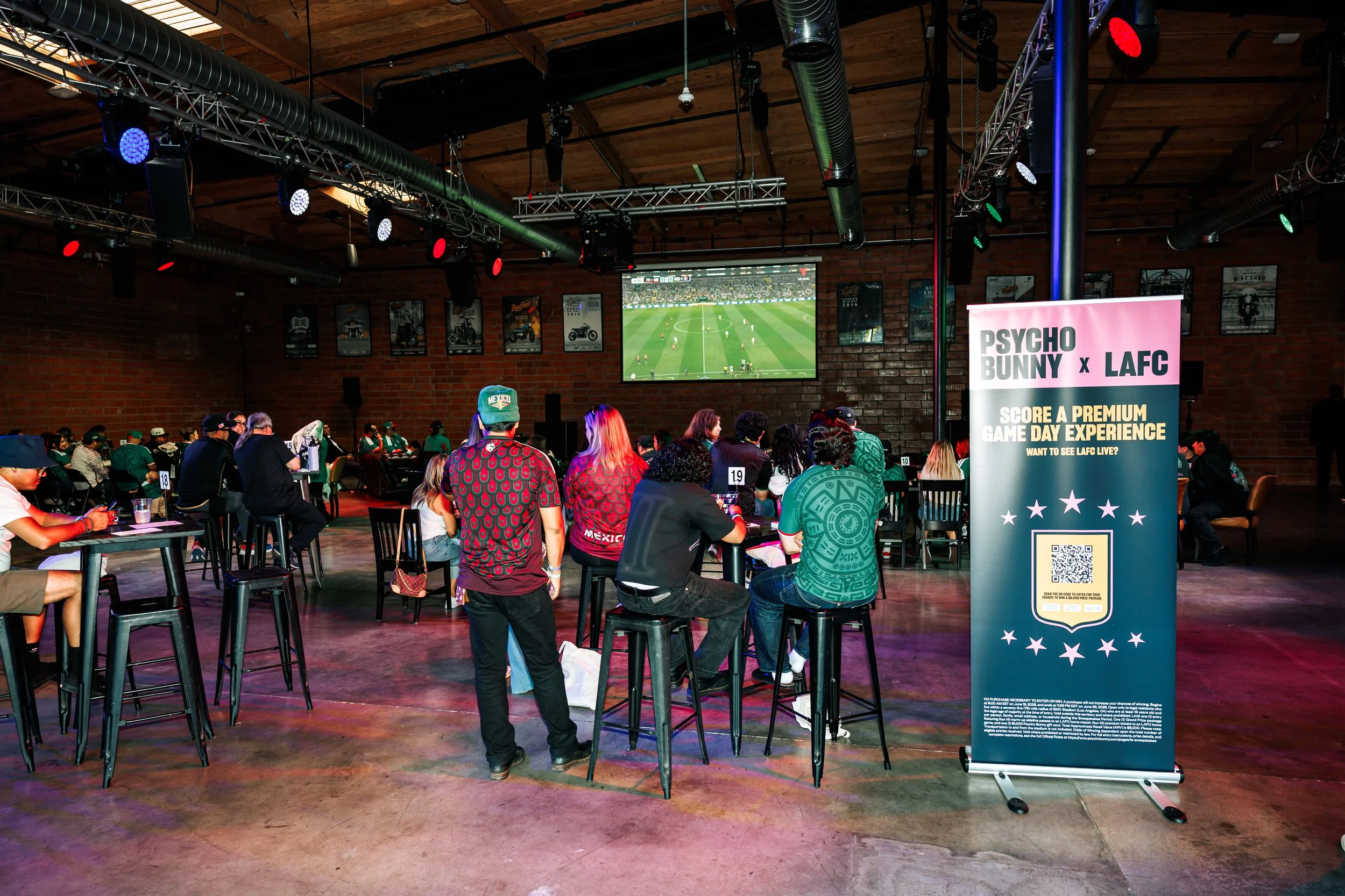 Distant image of soccer fans enjoying a watch party event at Bike Shed Moto LA. Large projection screens are in the background playing the soccer game