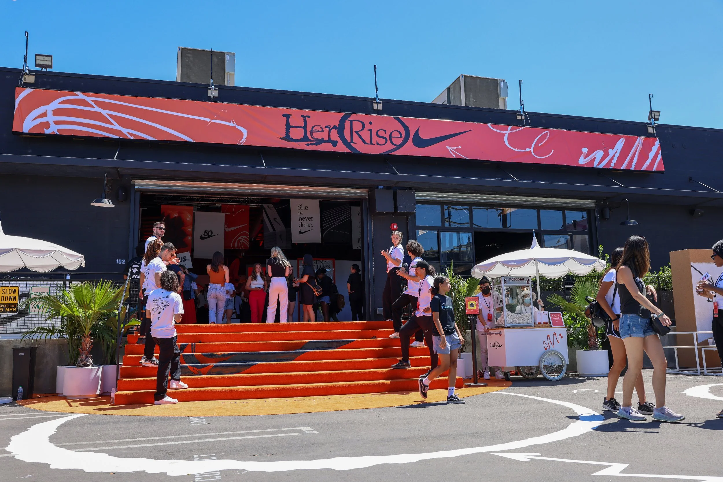 People gathering outside a Nike store with orange steps, a pink and black sign reading 'Her Rise,' and a popcorn cart.