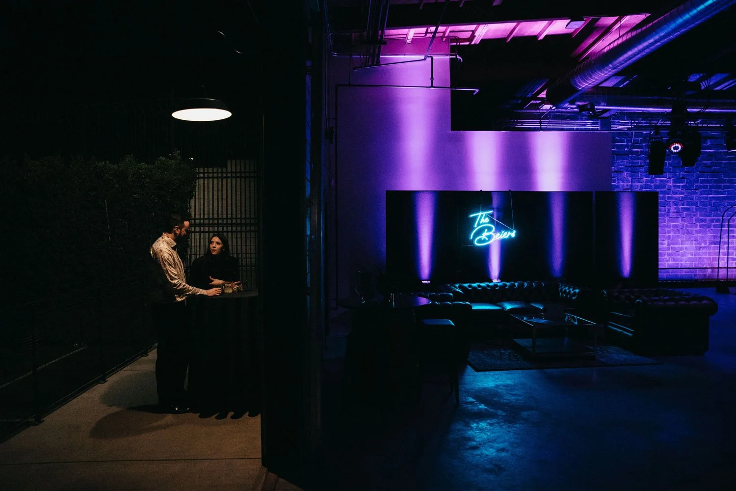 A dimly lit lounge or bar area with purple and blue lighting. A neon sign reads 'The Beat' on a wall with brick accents. Two people are standing near a small counter or bar on the left side, engaged in conversation.