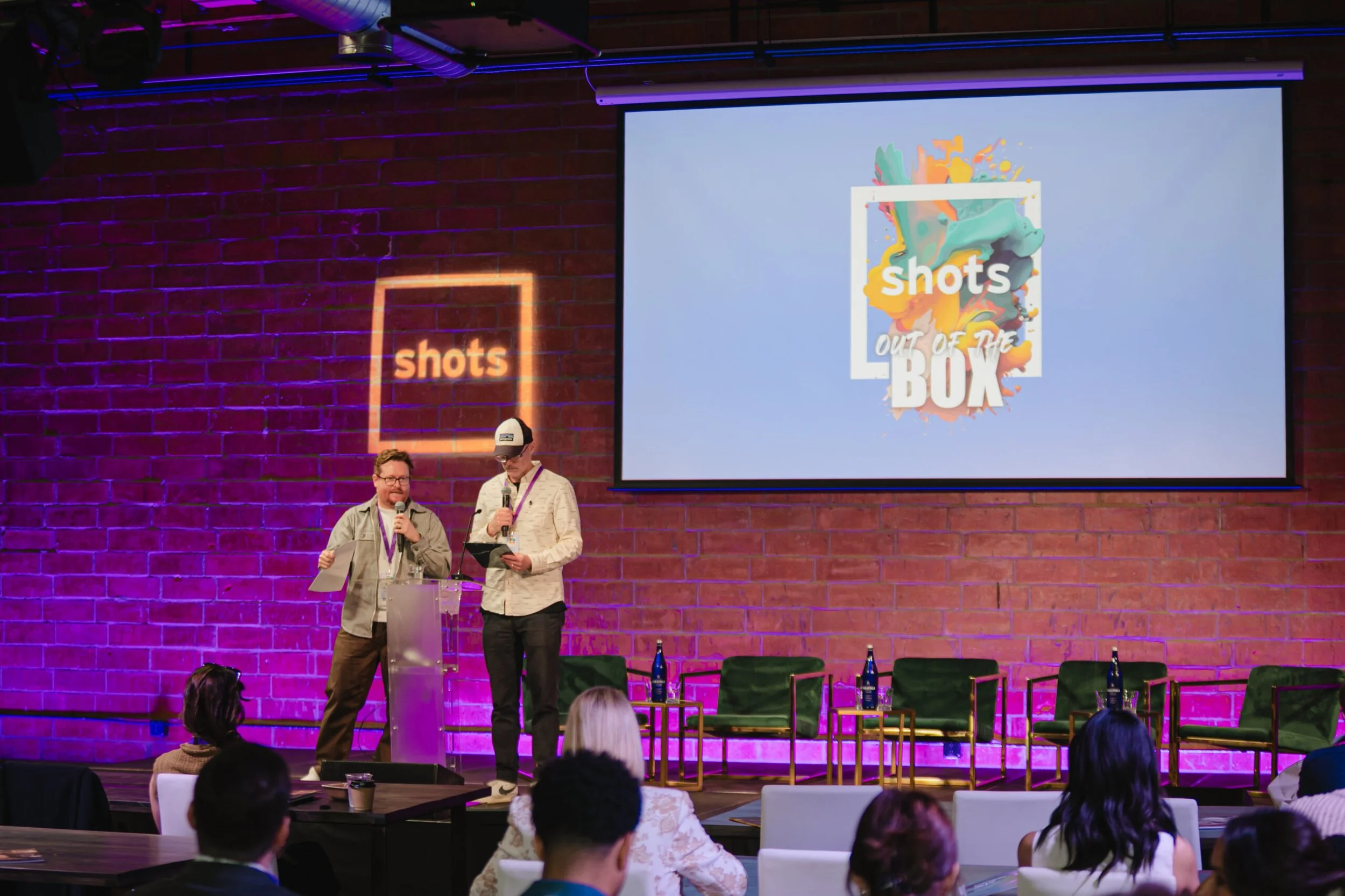Two men standing on stage in discussion during an event at Bike Shed Moto LA in front of large screen with a logo displayed on it