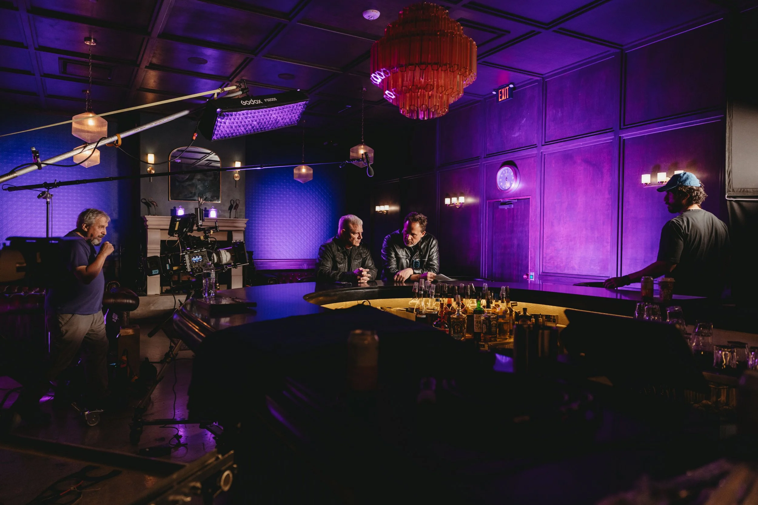 Dim indoor scene of two men talking a bar illuminated by some purple lighting. Filming of them chatting is taking place