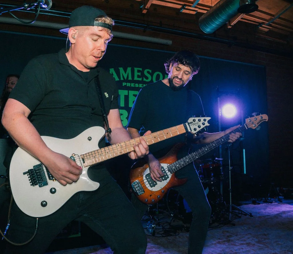 Two band members playing guitar and bass in front of signs at an event at Bike Shed Moto LA