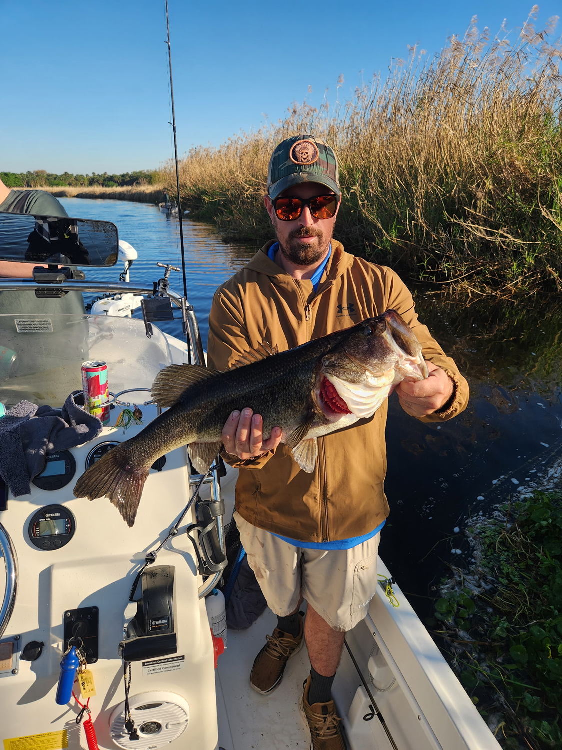 Man holding a large fish on a boat in a river with tall grass and blue sky in the background.