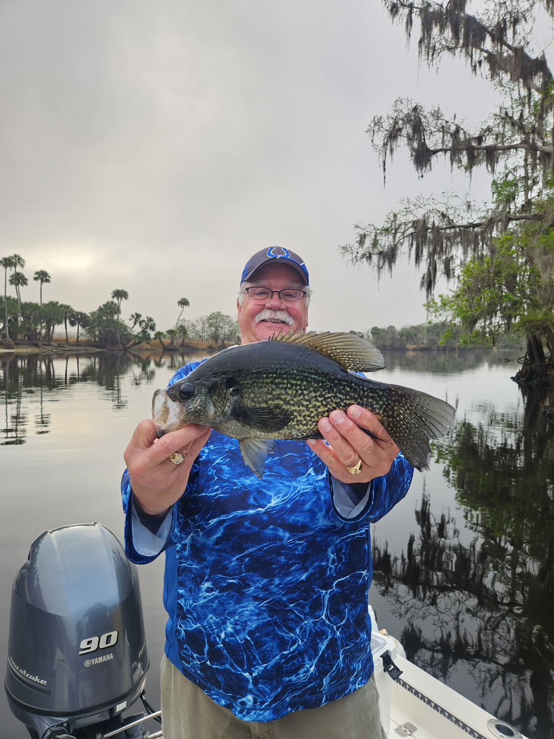 A man with a blue fishing shirt and cap holding a large fish seen on a boat during cloudy weather, with a river and trees in the background.
