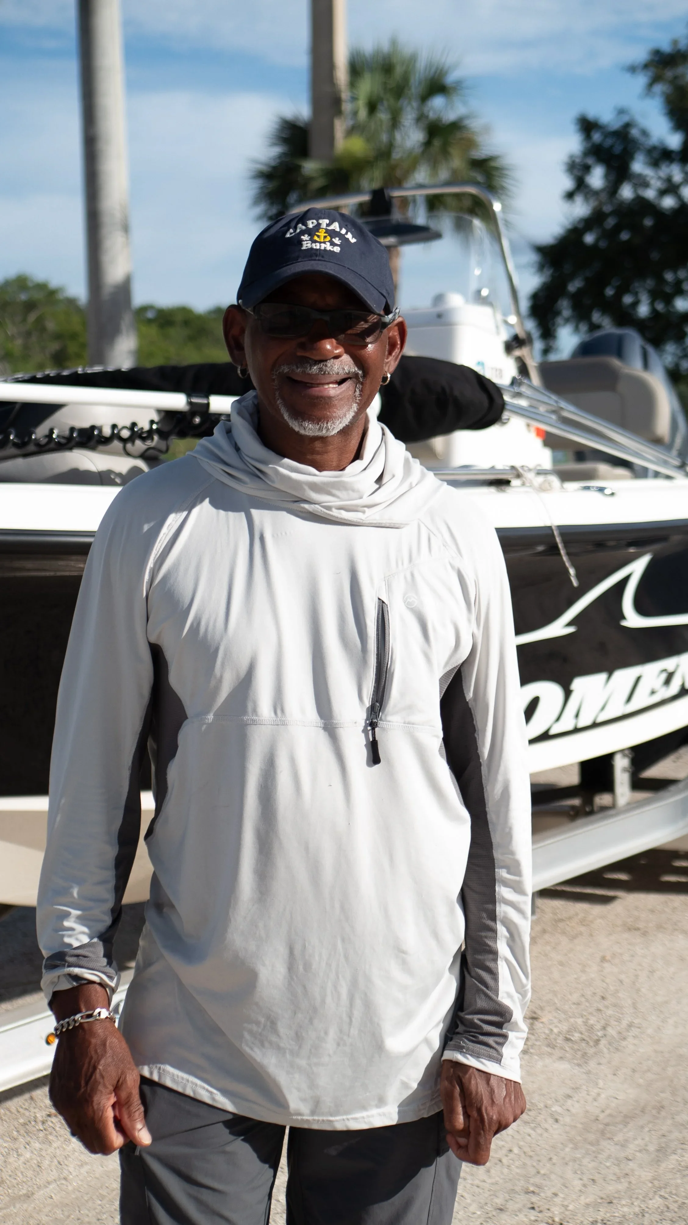 Smiling older man wearing sunglasses, a navy blue cap with 'Captain Barke' written on it, and a light-colored outdoor jacket, standing in front of a boat.