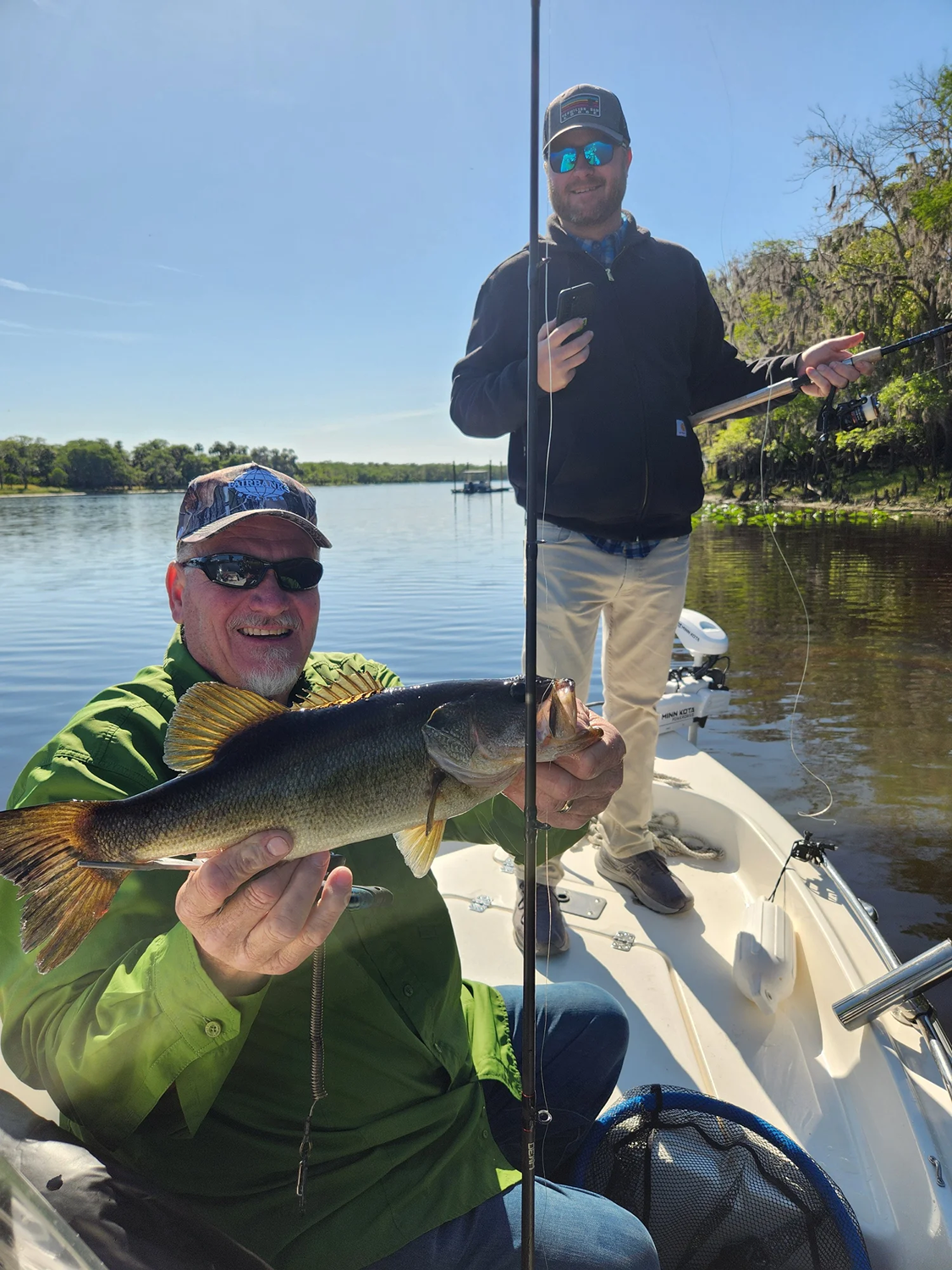 Two men on a boat by a calm river, one holding a large fish, with the other smiling and holding a fishing rod.