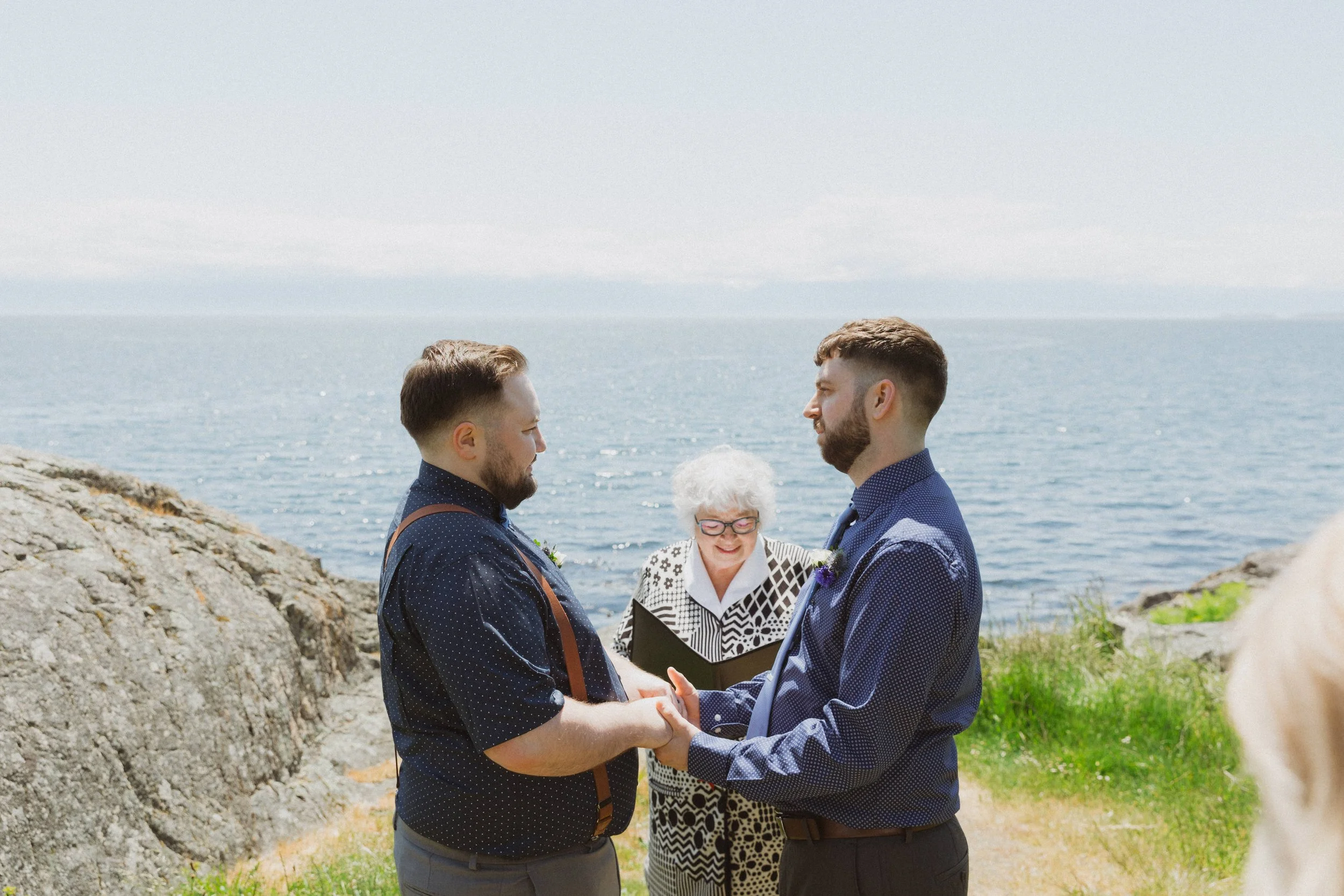 An officiant stands between two grooms during an intimate oceanfront elopement ceremony in Victoria, BC.