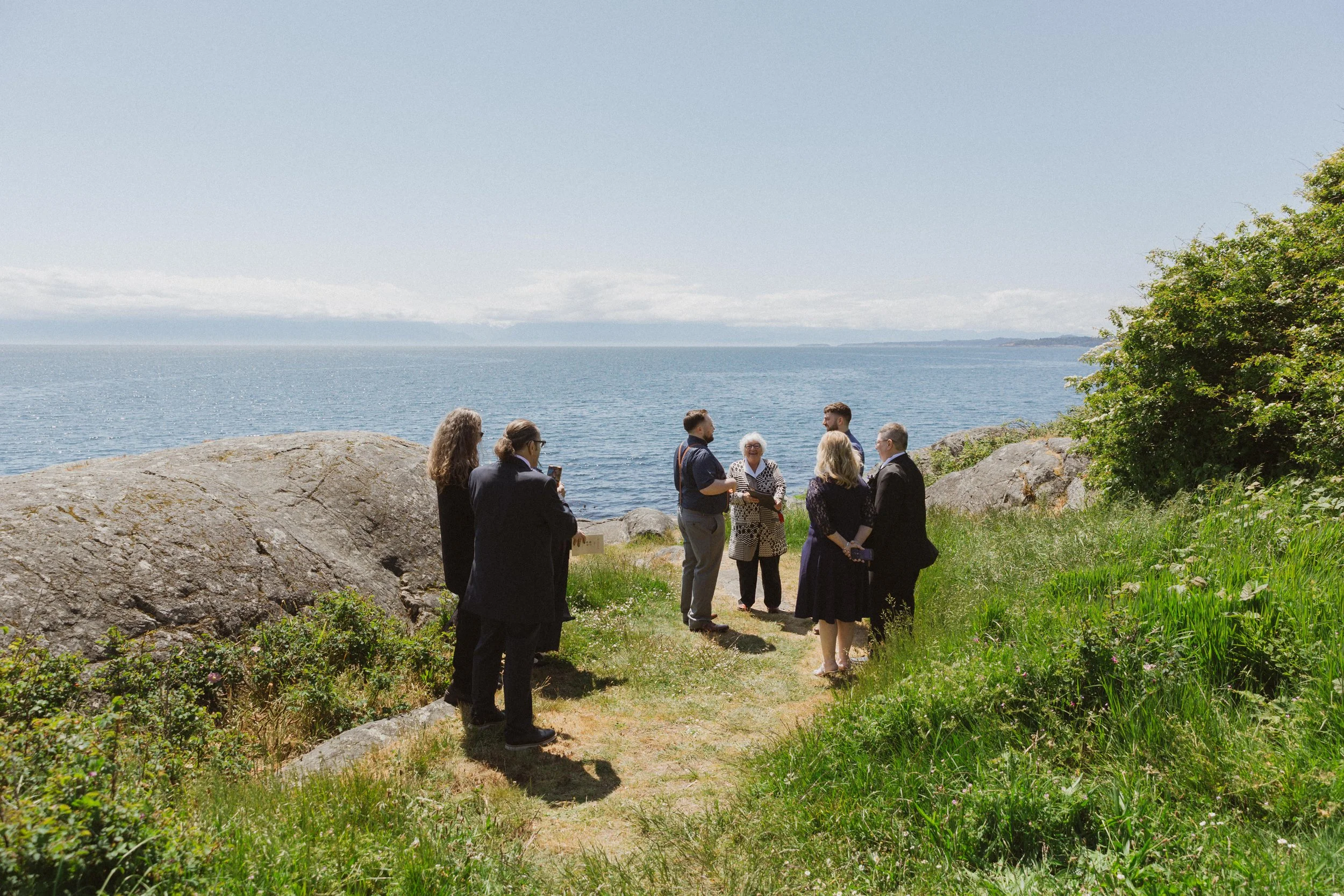 Two grooms stand facing each other during an intimate oceanfront elopement ceremony in Victoria, BC while their parents and officiant stand nearby.