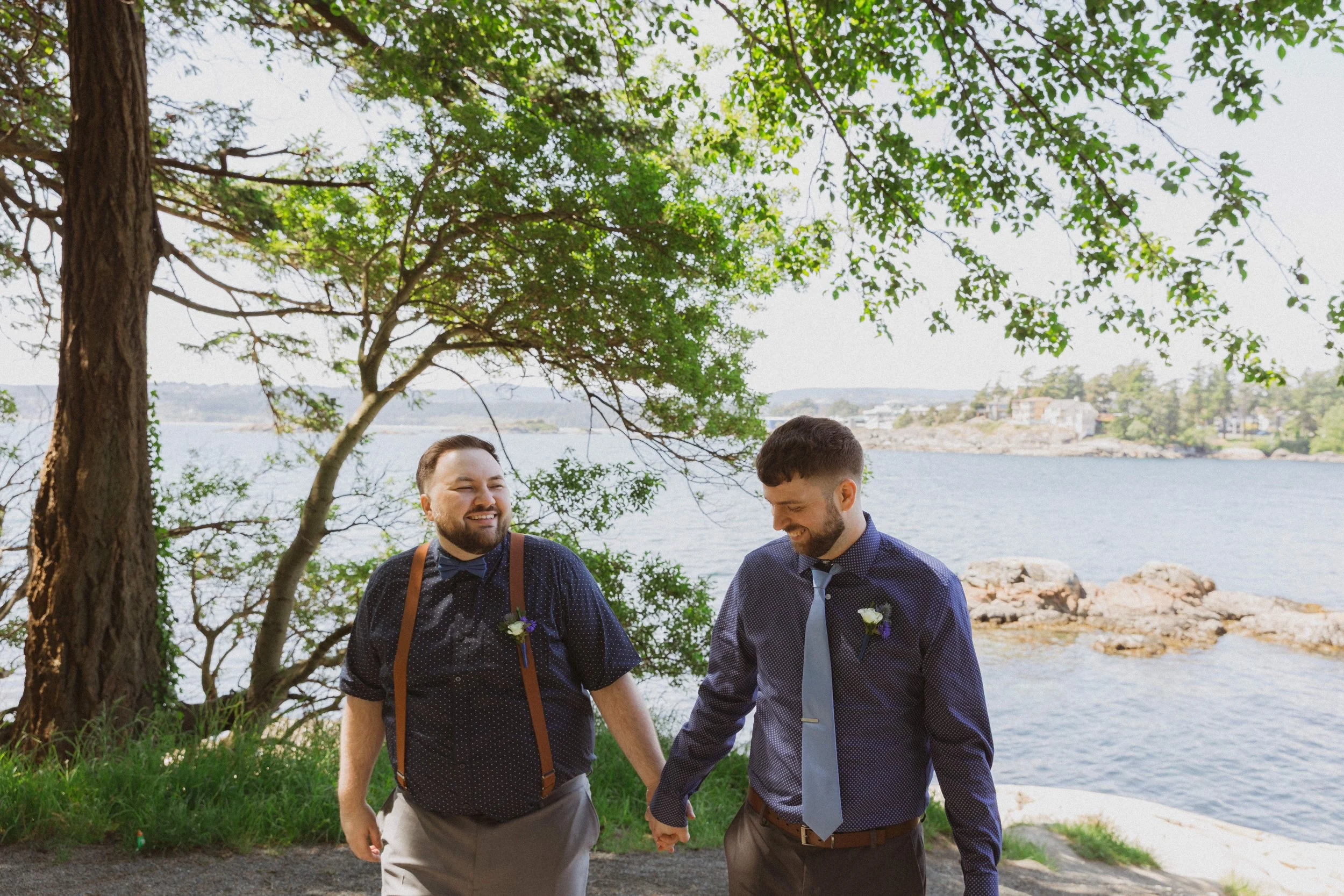 The newly married grooms walk hand in hand smiling with the ocean and a large tree behind them in Victoria, BC.