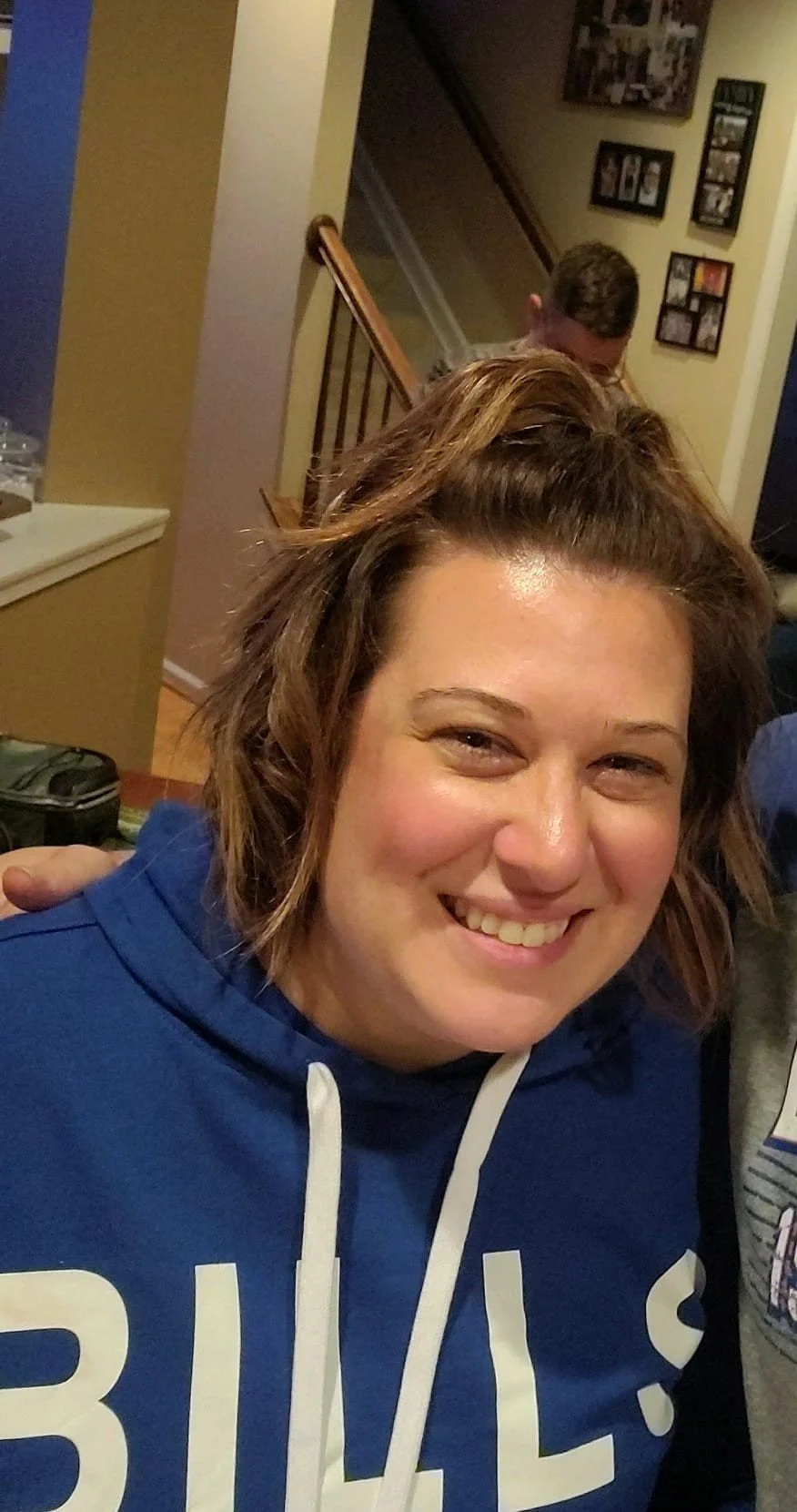 Megan is wearing a blue hoodie, in a home interior with a staircase and picture frames on the wall in the background.