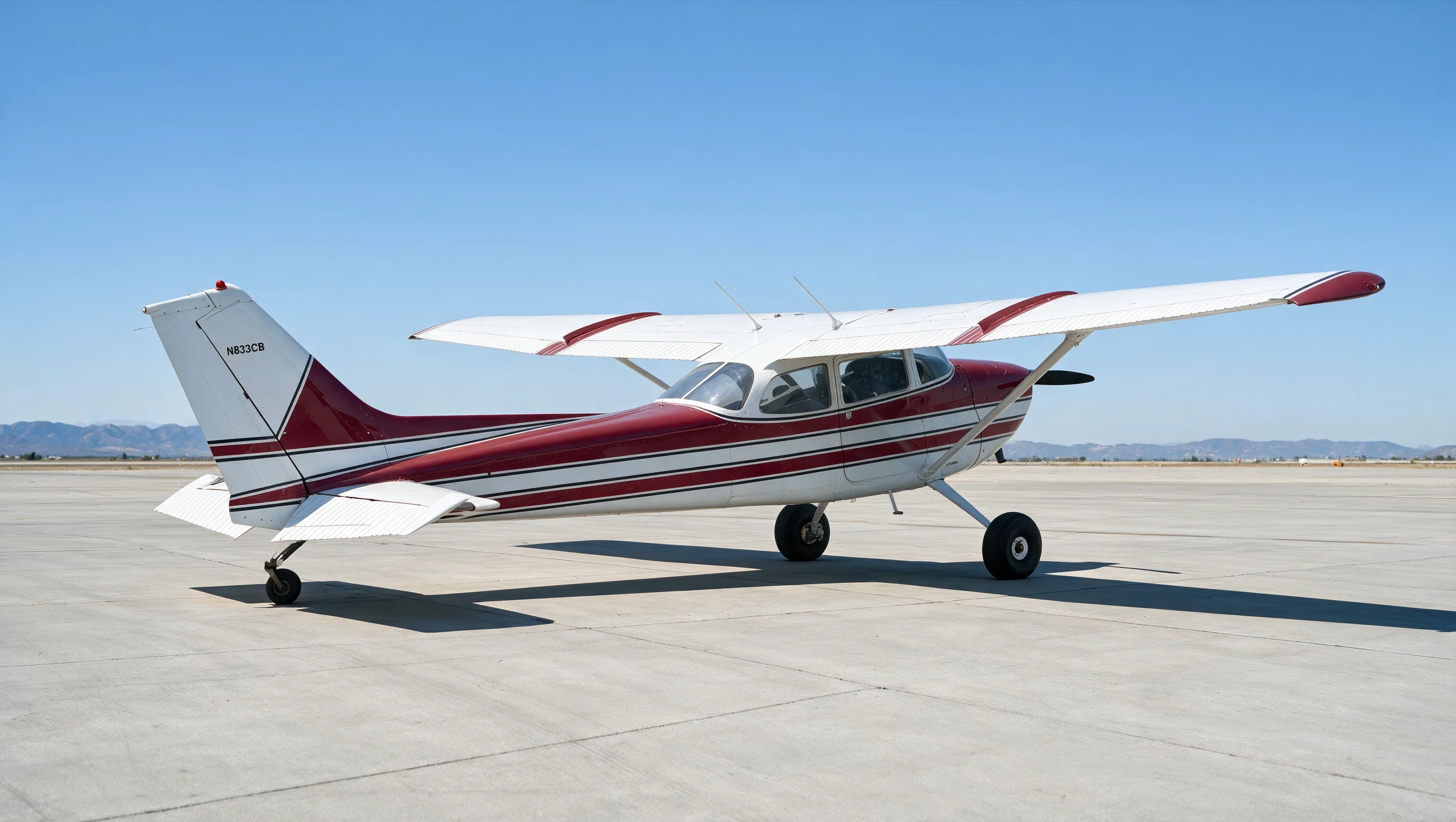 A small white airplane with red and black stripes is parked on a tarmac under a clear blue sky, with mountains in the background.