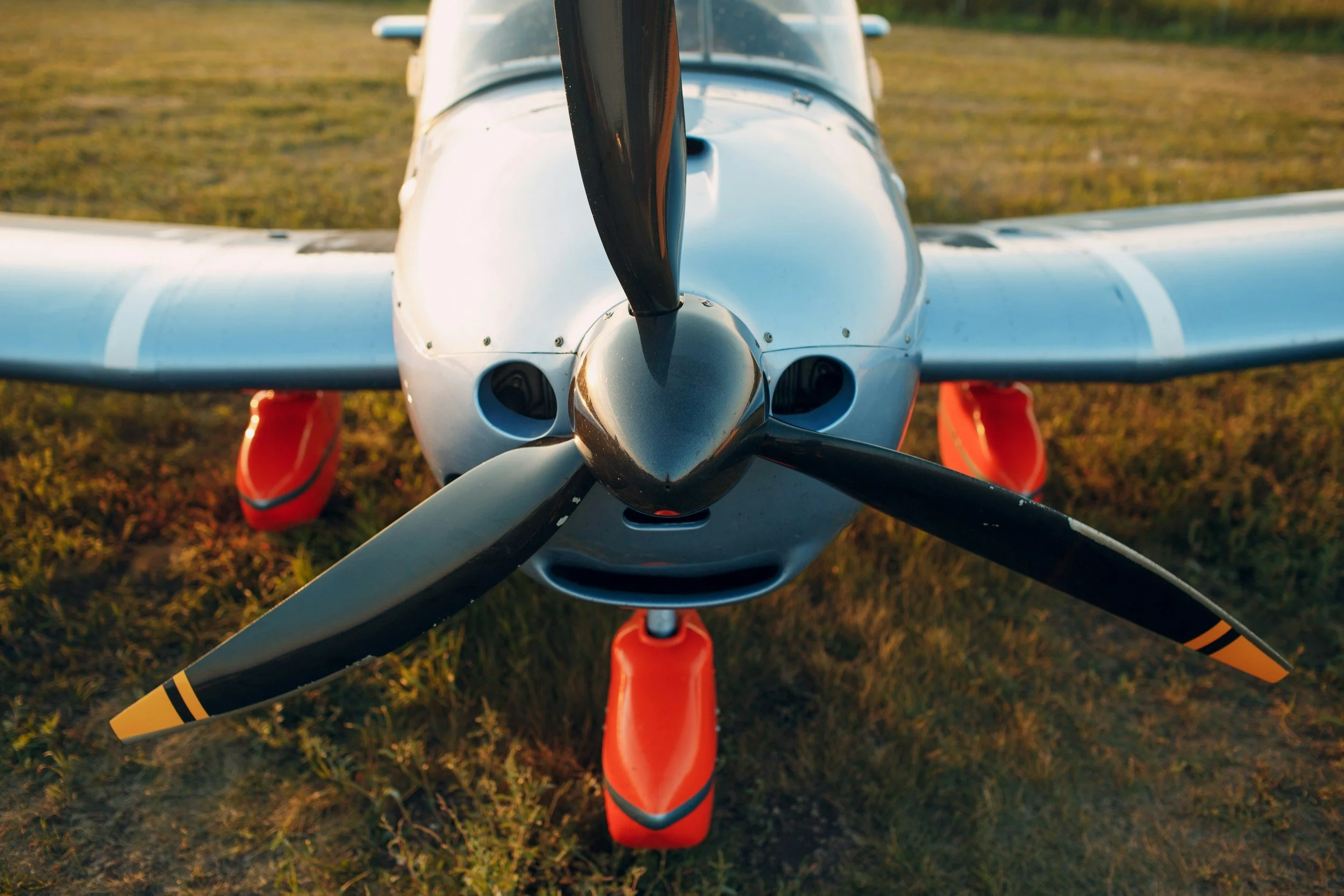 Front view of a small white airplane with a black propeller, orange landing gear, and a grassy field background.