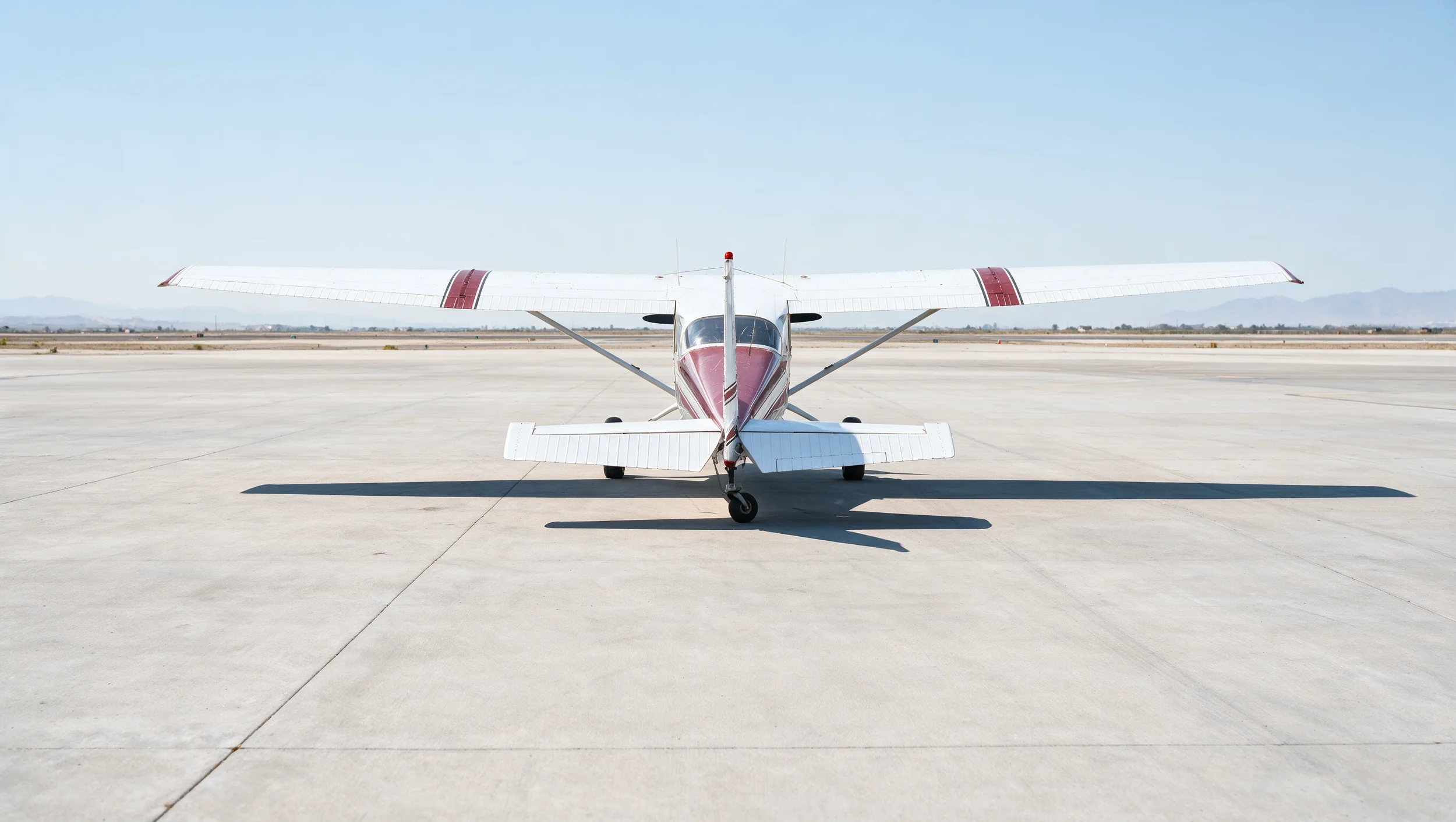 Small white and red airplane parked on a wide concrete tarmac at an airport with mountains in the distance and a clear sky overhead.