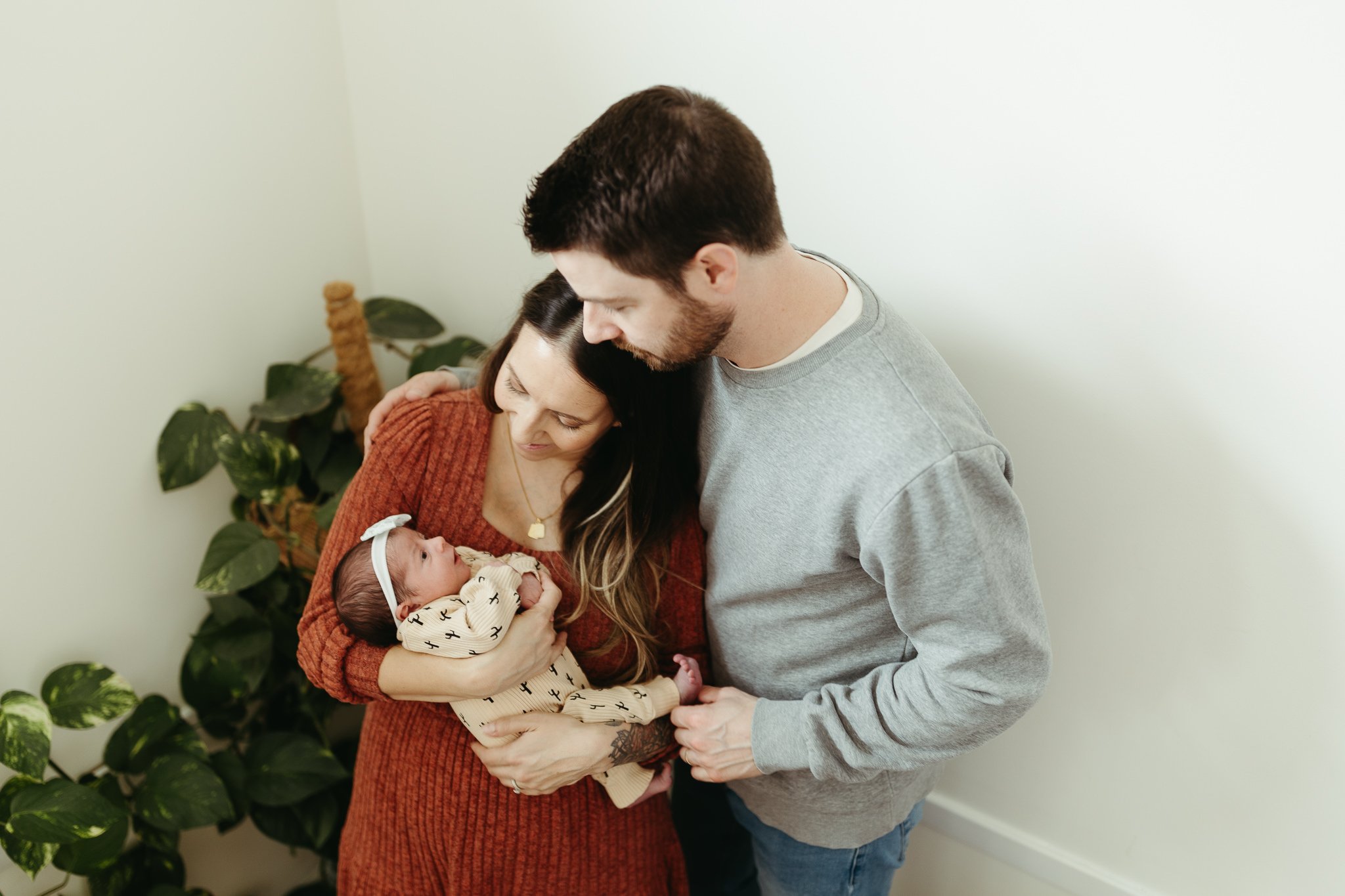 New mom in red dress and dad in grey sweater hold their newborn daughter at their home in Calgary