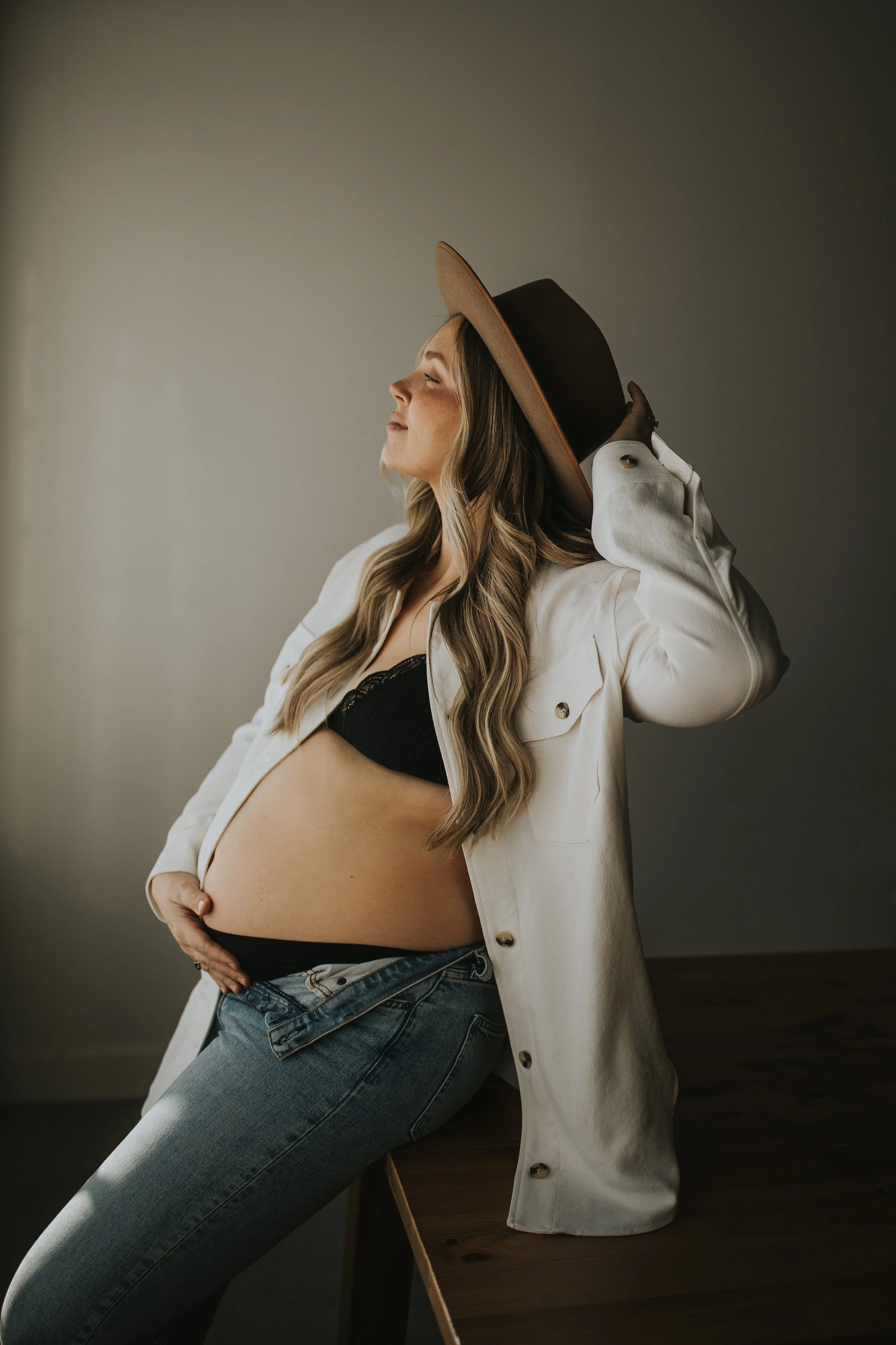 Pregnant woman wearing a black bra, white shirt, jeans, and brown hat, posed sideways, smiling, with hand on her belly.