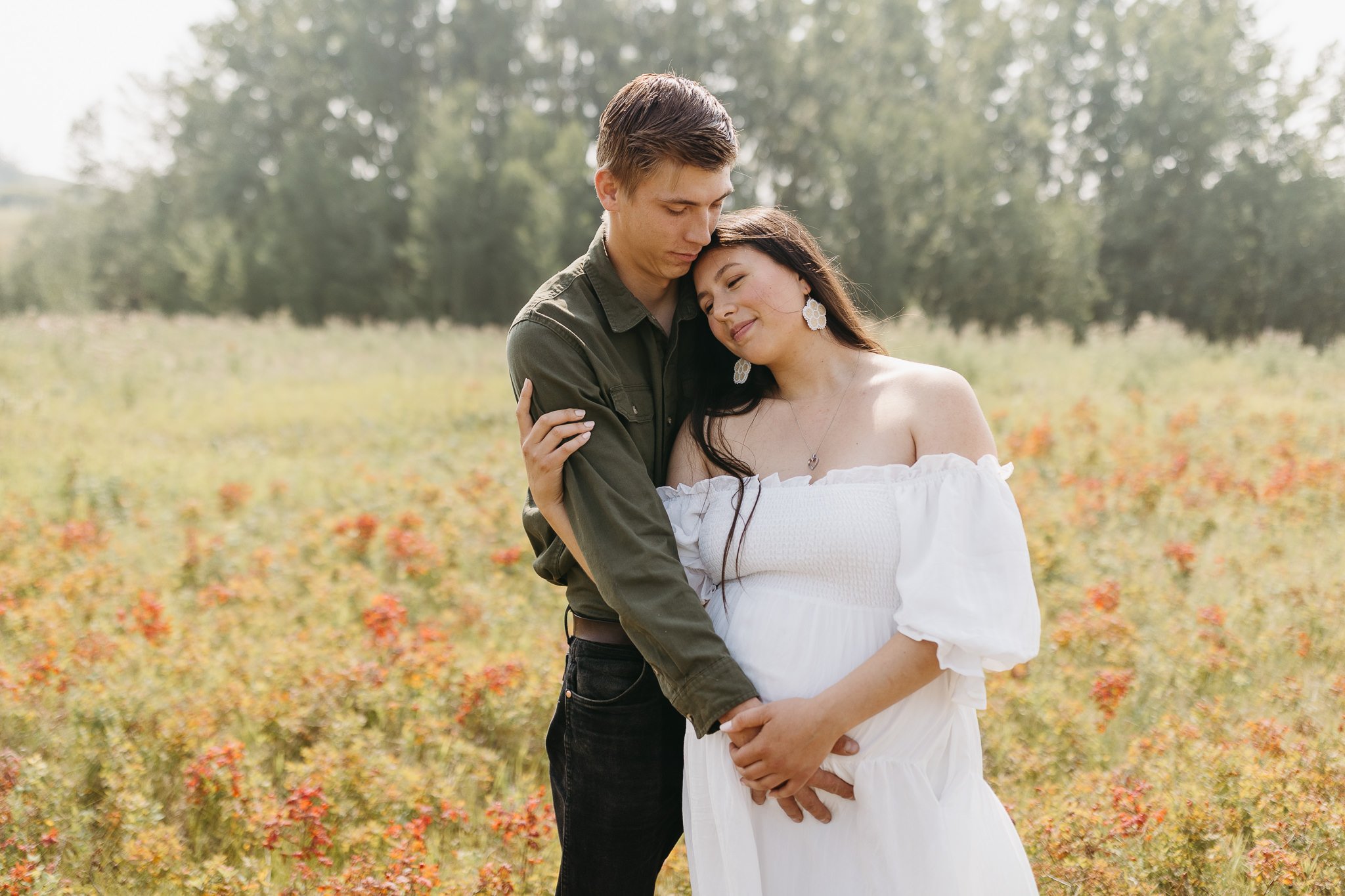 Pregnant woman in white dress stands in meadow with red wildflowers during her fall maternity session in Calgary
