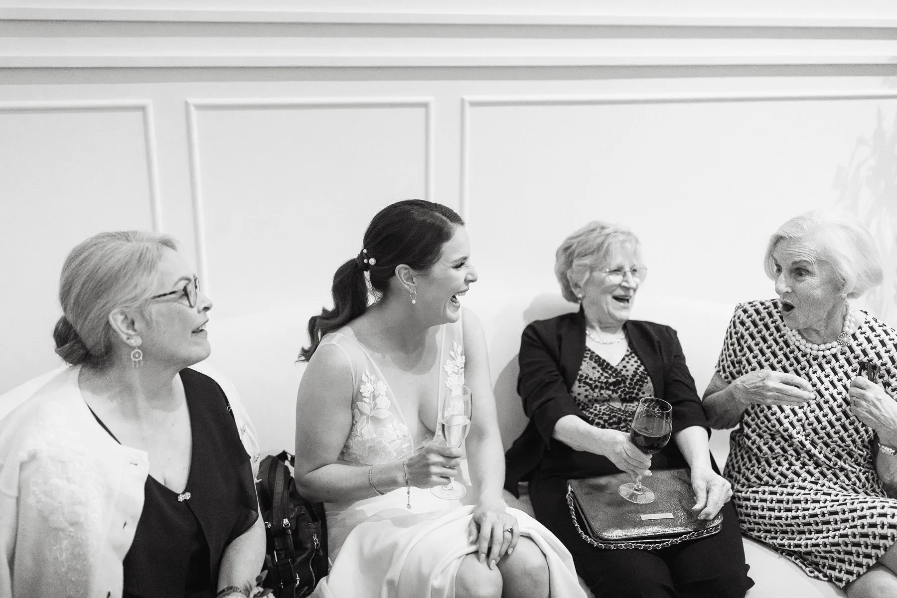 A black and white photo of four women sitting on a couch, laughing and holding drinks, with one wearing a wedding dress at Venue 308 in Calgary, Alberta