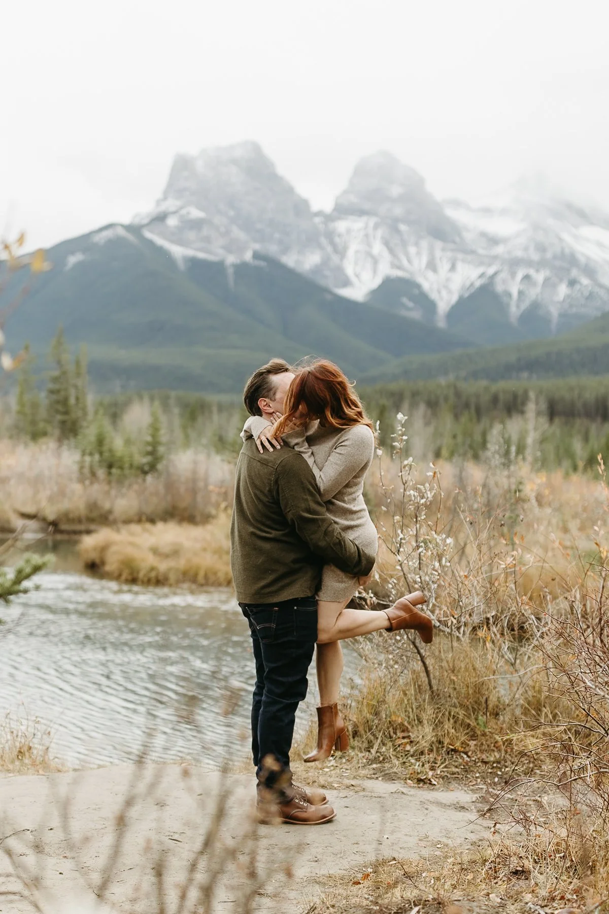 Couple embracing during their engagement session near a river with snow-capped Three Sisters mountains in the background in Canmore, Alberta