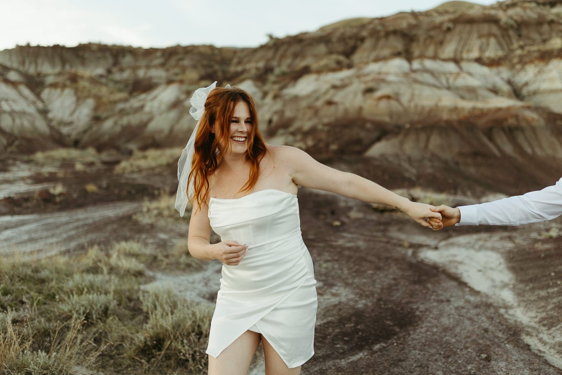 Laughing bride in white dress and veil holding groom's hand in rocky outdoor setting in Drumheller, Alberta