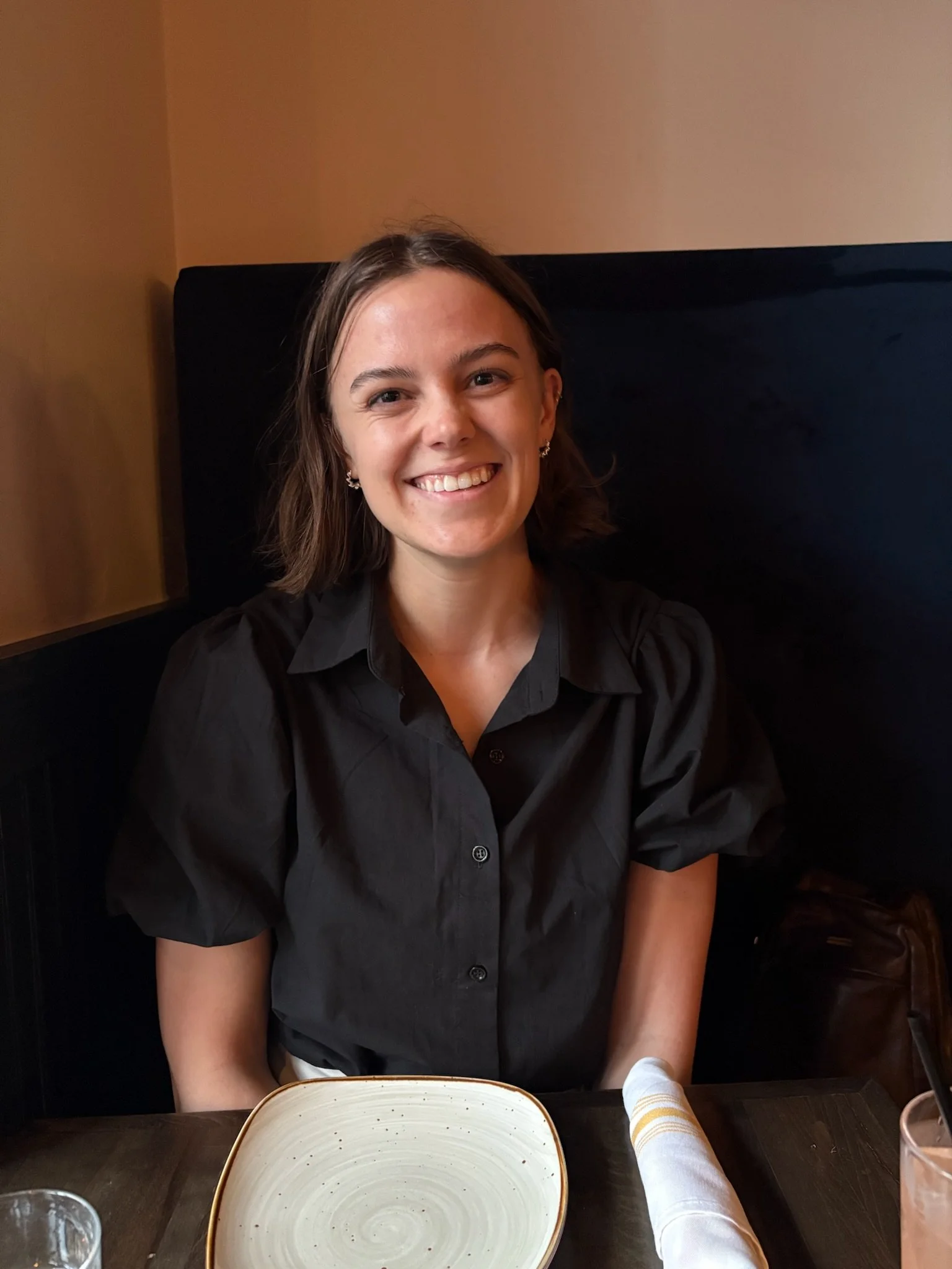 Smiling woman with short brown hair, wearing a black shirt, sitting at a restaurant table with an empty plate and hand towel in the foreground.