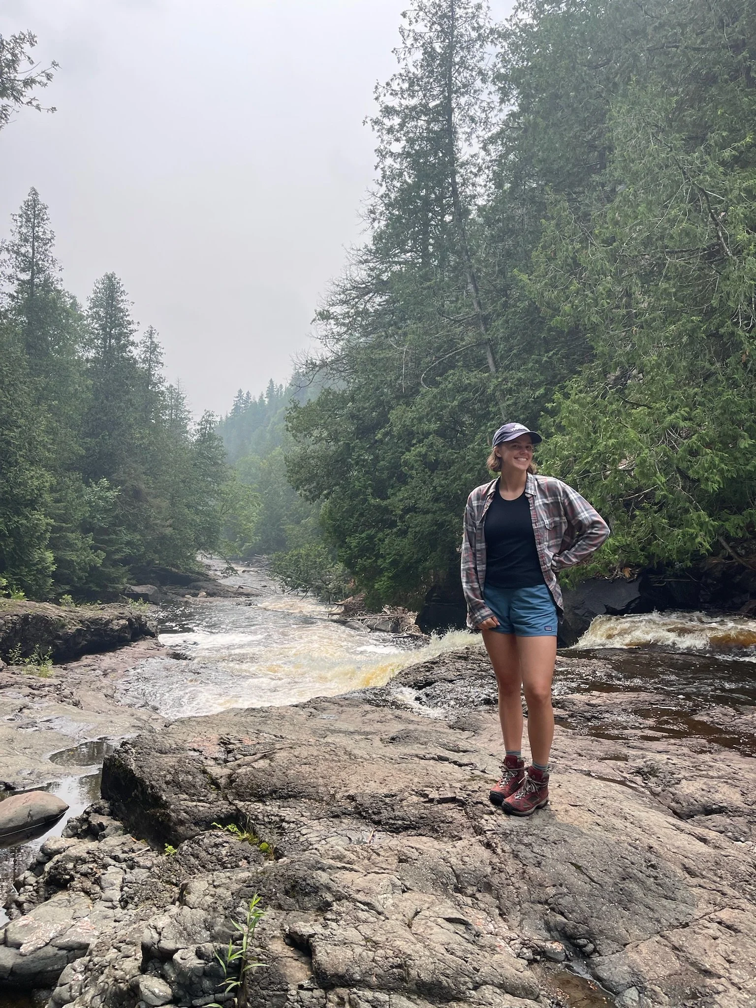 A woman smiling and standing on rocky terrain by a flowing river in a green forest with tall trees, under an overcast sky.