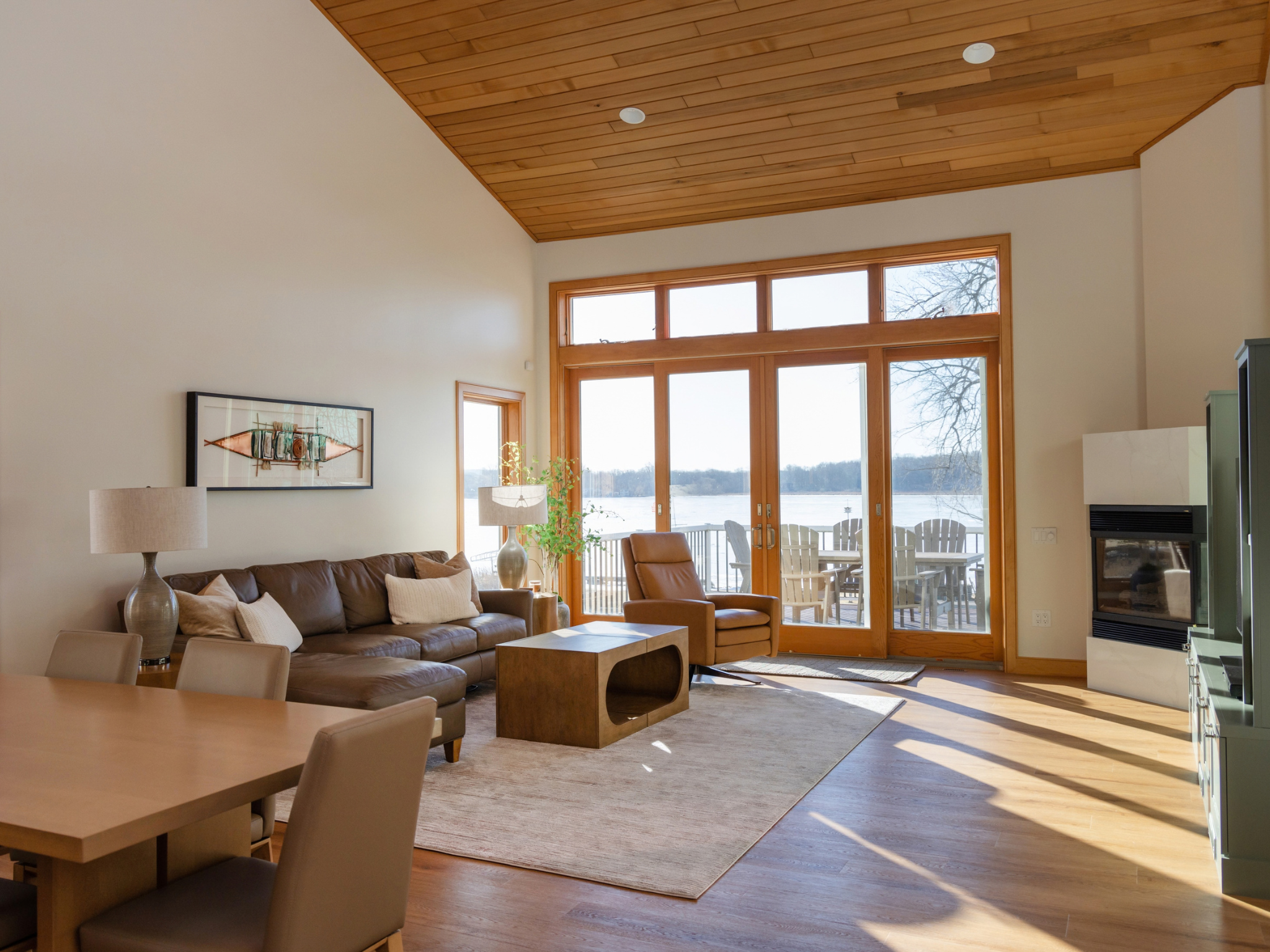 Custom home design featuring a sunlit living room with a vaulted wood ceiling, leather sofa, dining table, fireplace, and tall windows overlooking a lake.