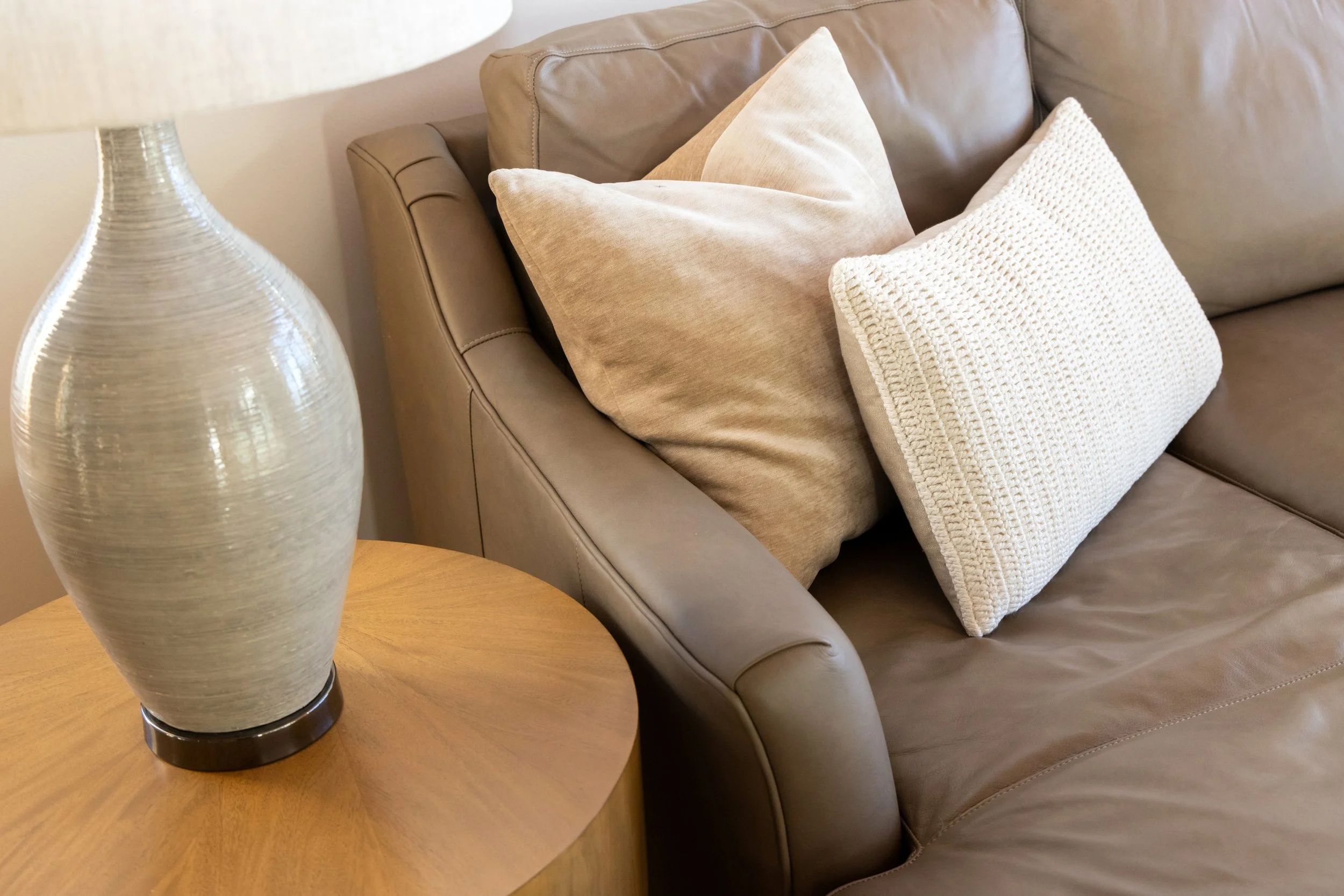 Custom home interior vignette with brown leather sofa, cream pillows, round wood side table, and textured lamp.
