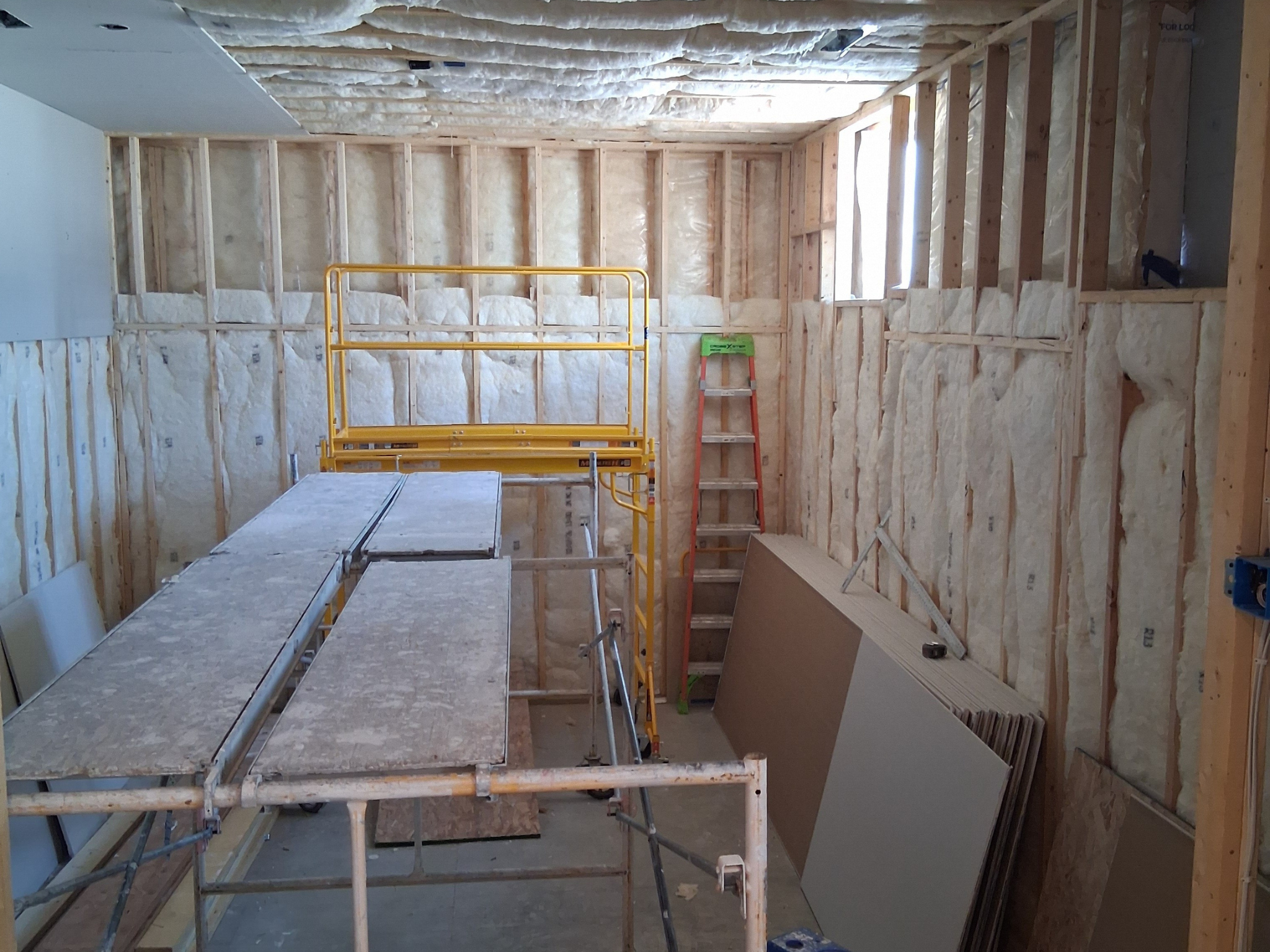 Custom home construction in progress with exposed wood framing, insulation, drywall panels, work platforms, and ladder.