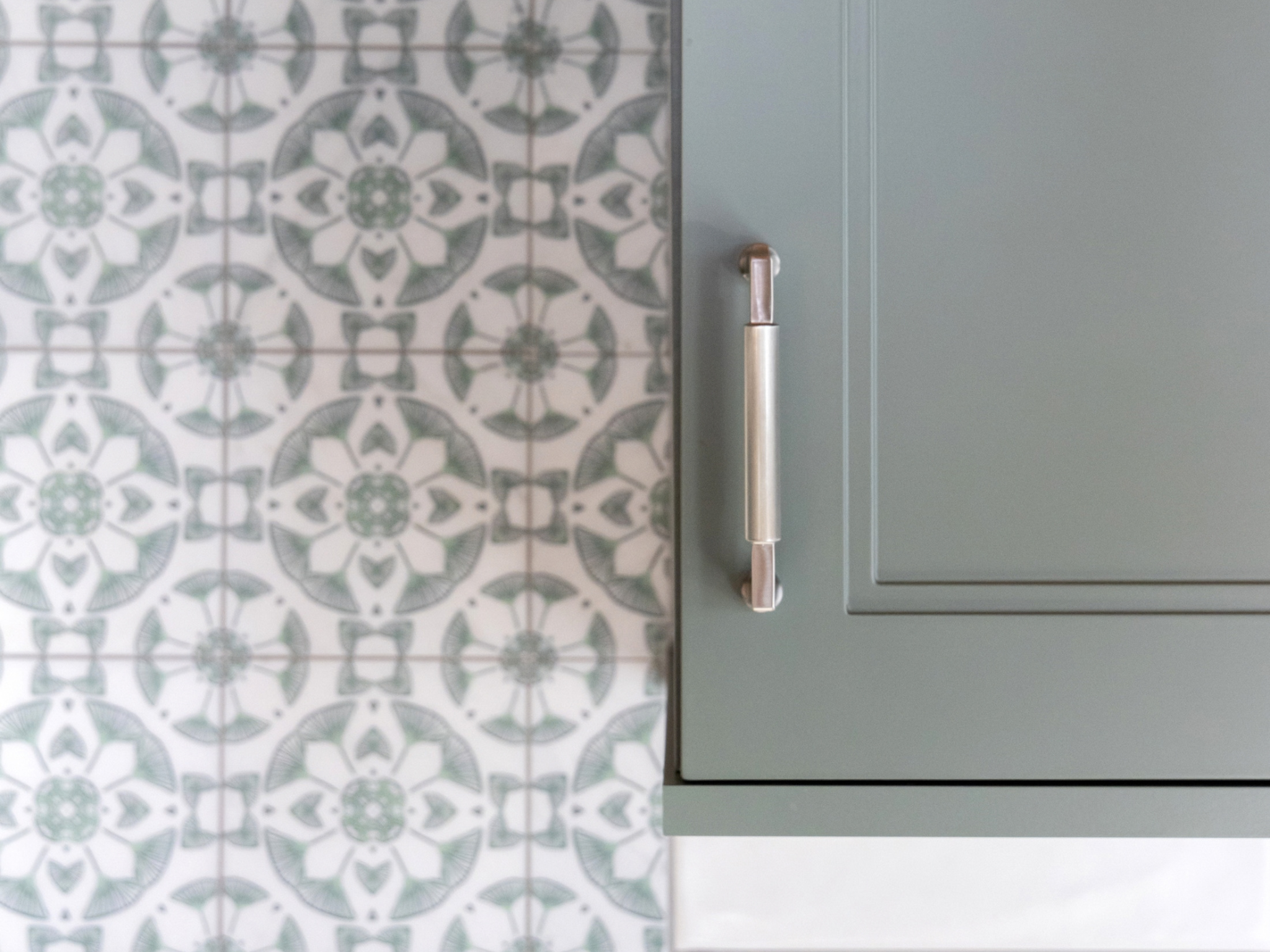 Custom home kitchen with muted green cabinets, slim metal handles, patterned tile backsplash, and white range hood in a calm, vintage-inspired design.