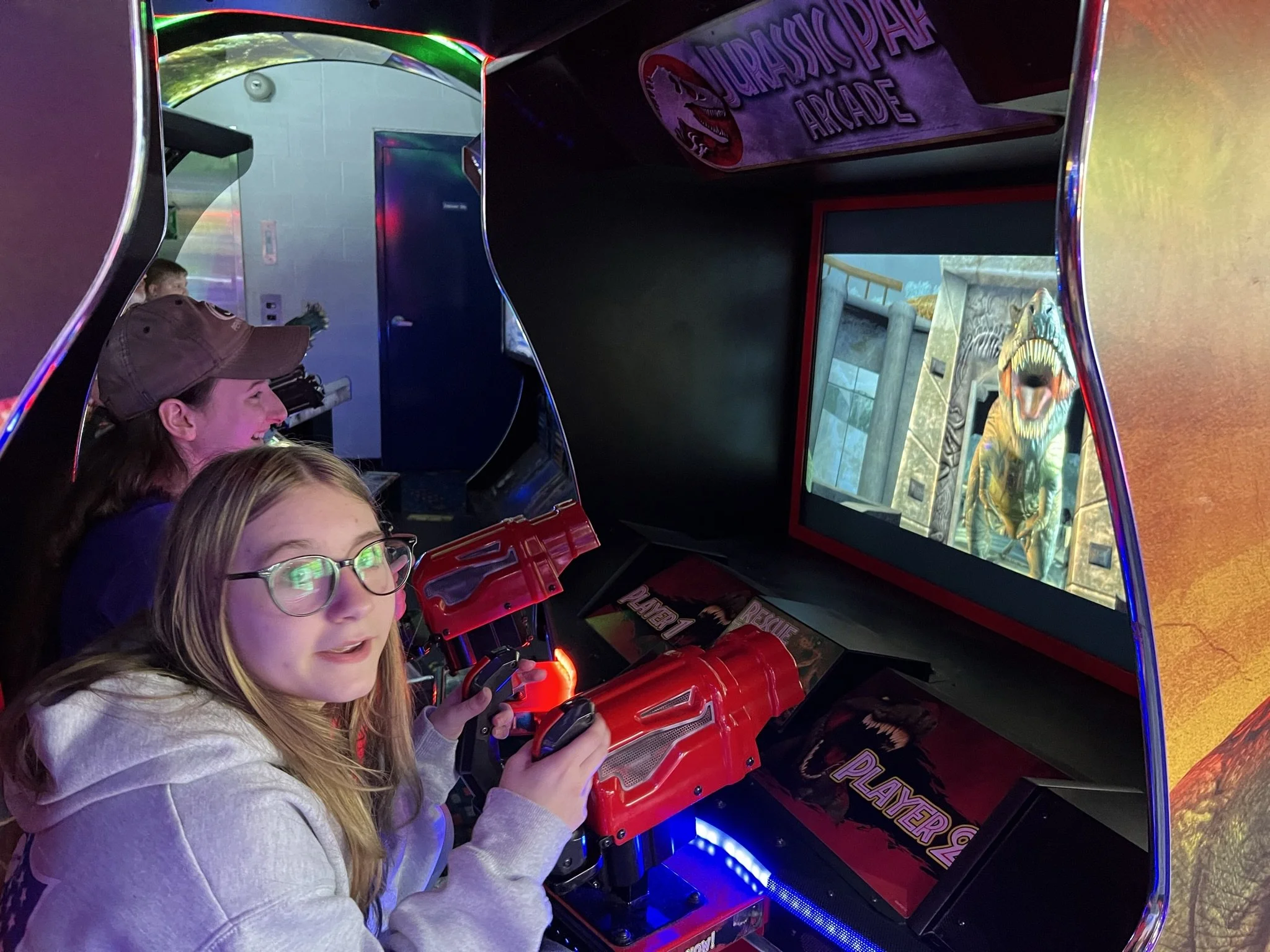Two children playing Jurassic Park arcade game, with a woman watching, in an arcade setting with colorful lighting.