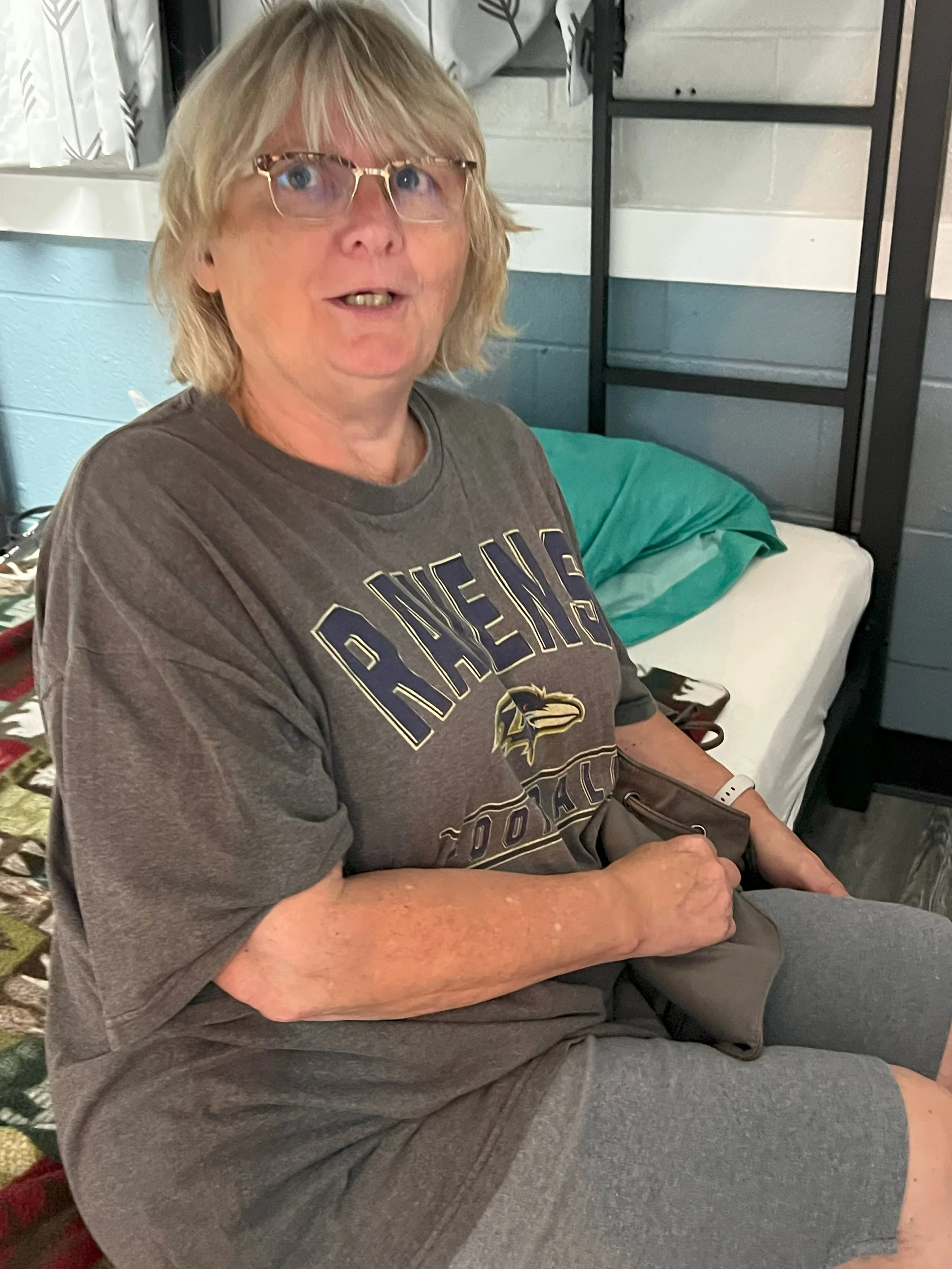 An older woman with blonde hair and glasses sitting on a bed, wearing a gray Ravens T-shirt and shorts, holding a small bag, with a teal pillow behind her and a bed with a quilted blanket next to her.