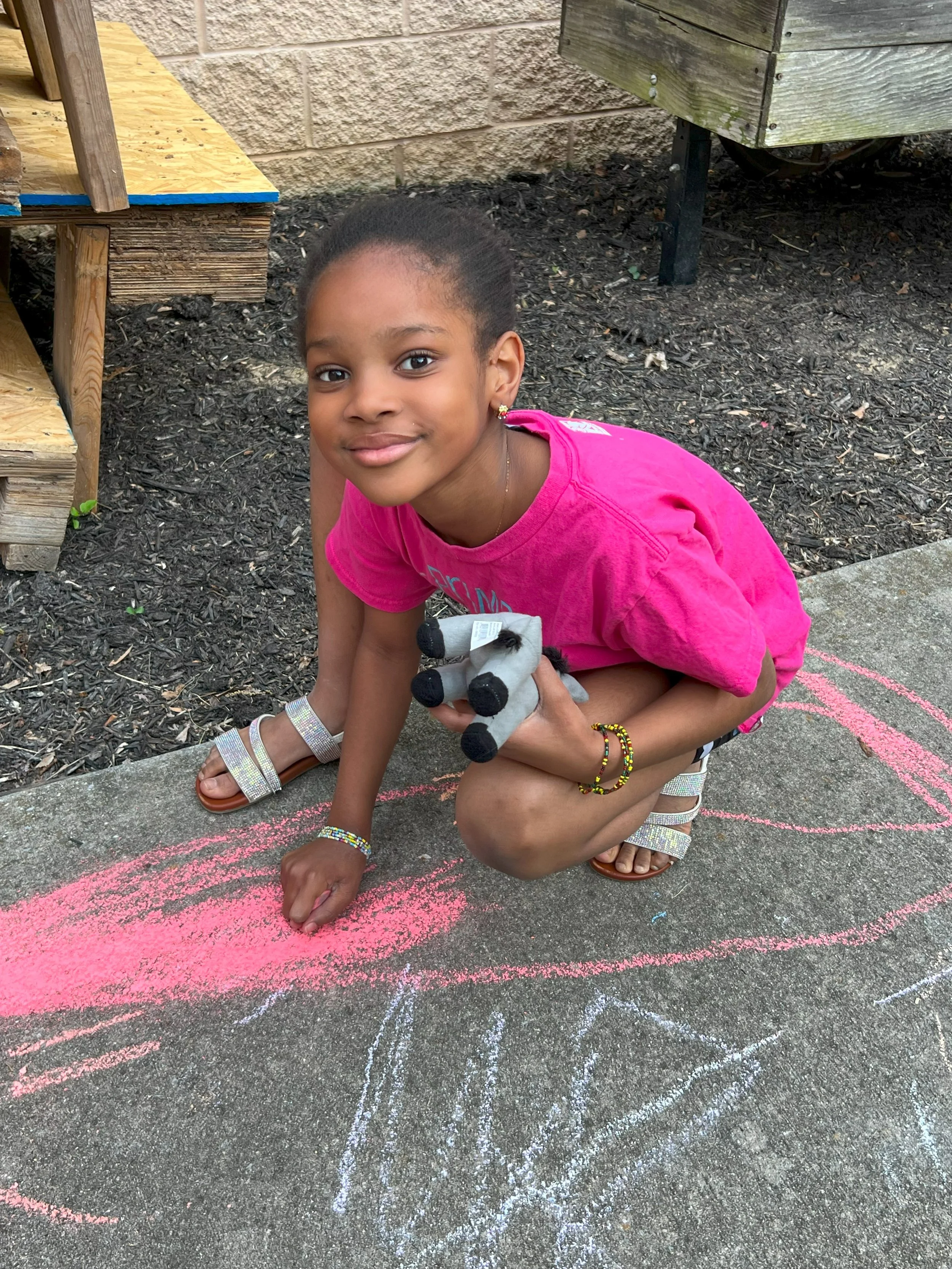 A young girl in a pink shirt drawing with sidewalk chalk on pavement, holding a stuffed animal, with a smile on her face.