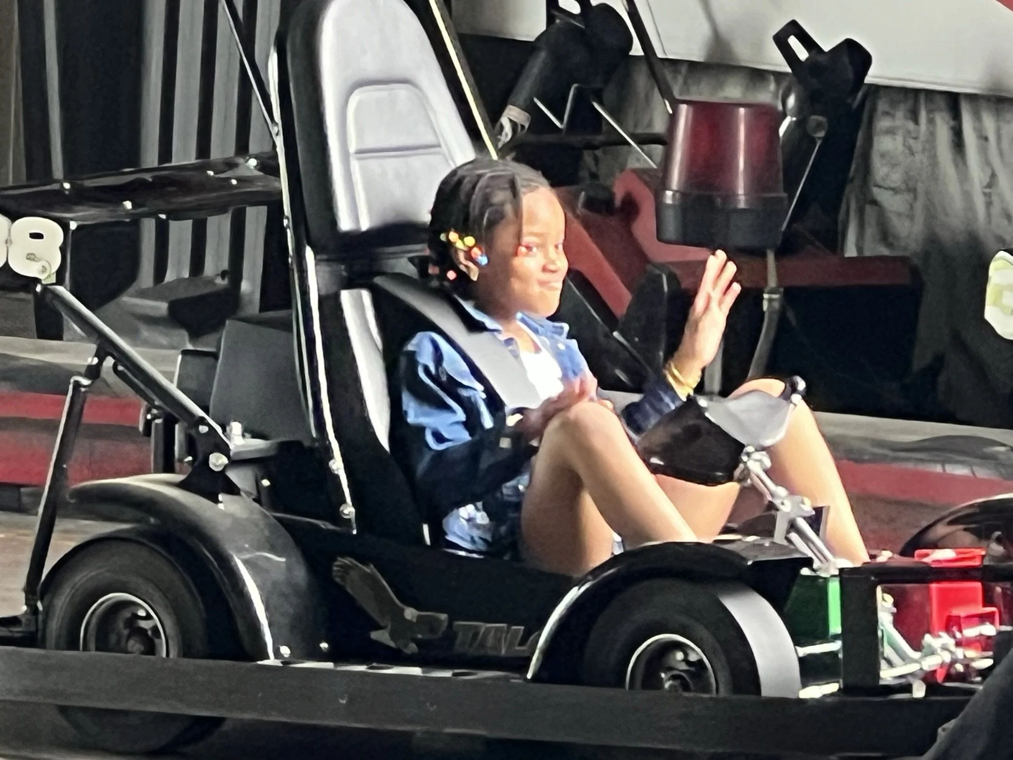 A child sitting in a go-kart, waving with a smile, and wearing a blue jacket and shorts, with braided hair decorated with colorful beads.