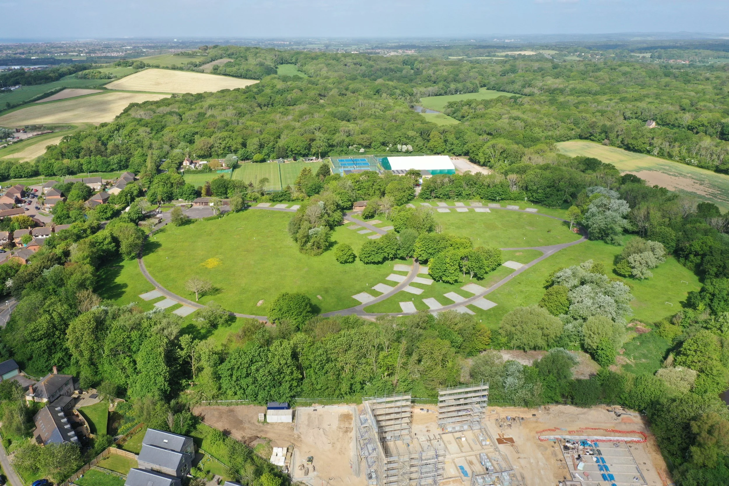 Aerial view of a park with green trees, open grassy areas, pathways, and tennis courts, surrounded by residential houses and construction site at the bottom, with farmland and wooded areas in the background.
