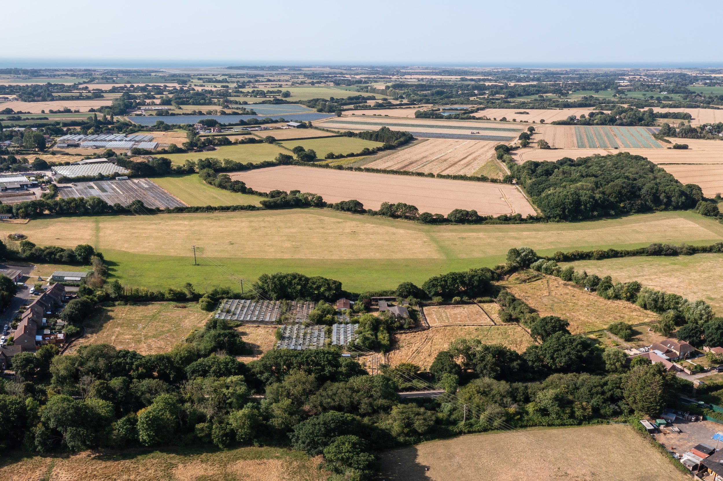 Aerial view of a rural landscape with fields, trees, and small houses extending to the ocean in the distance, under a clear sky.
