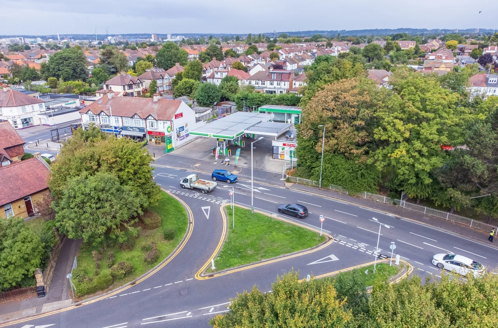 Aerial view of a gas station with cars parked and driving on a nearby street, surrounded by trees and residential houses in a suburban neighborhood.