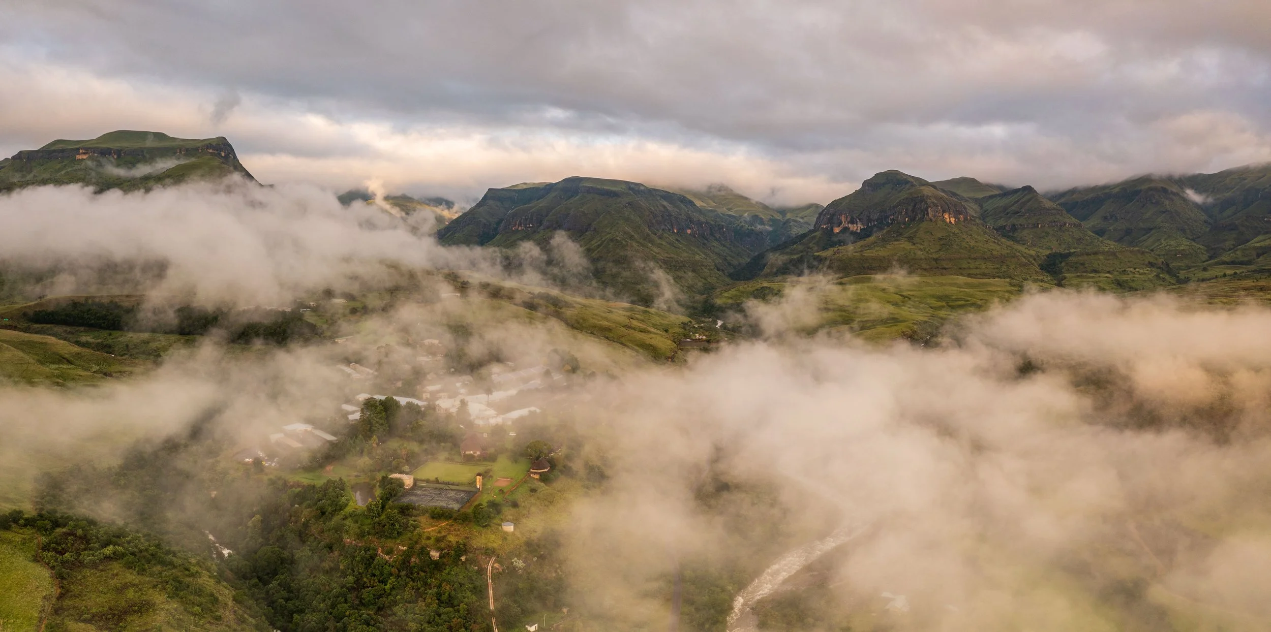A mountainous landscape with green hills and cliffs, partially covered by fog and clouds, with a small village visible at the base of the hills.