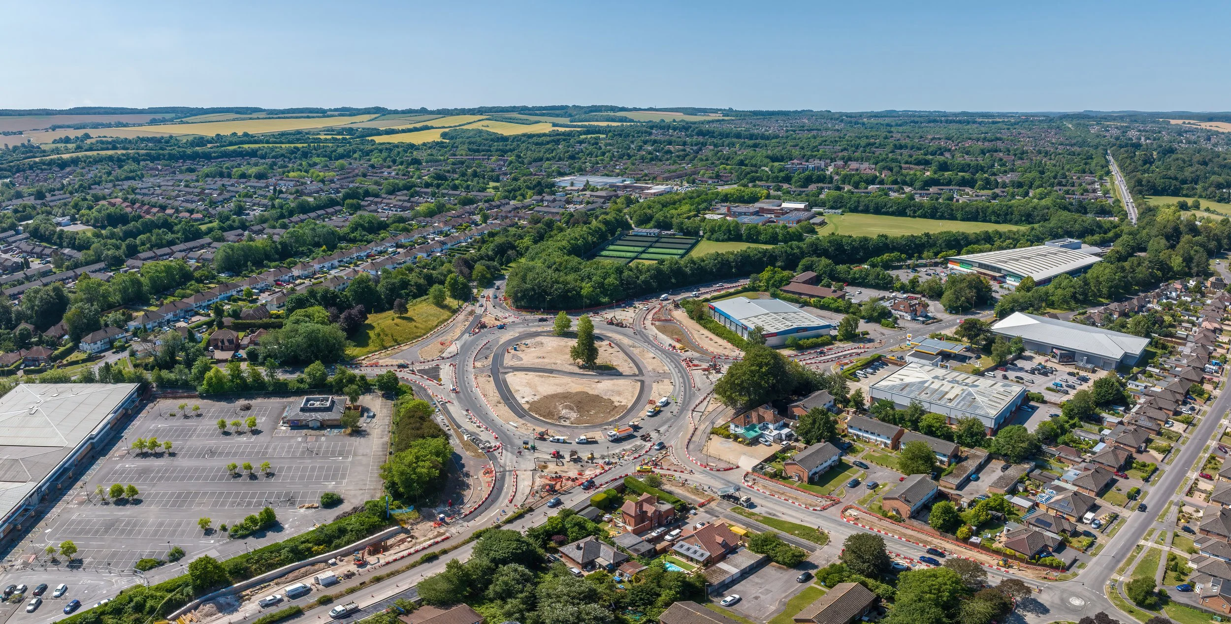 Aerial view of a roundabout under construction in a suburban area, surrounded by houses, commercial buildings, and green spaces, with a parking lot in the foreground and rolling hills in the background.