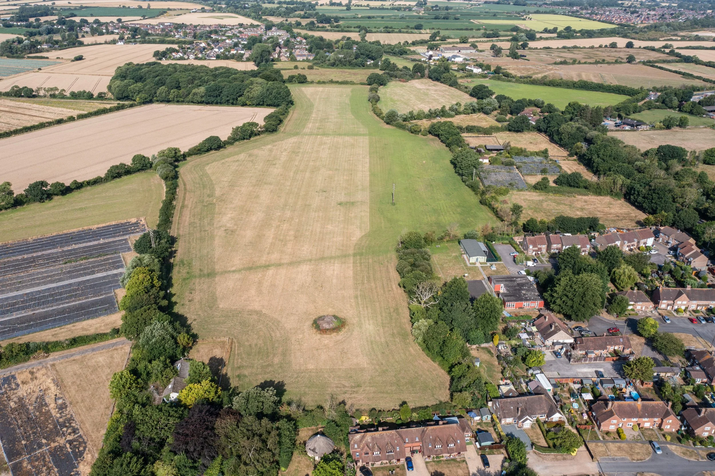 Aerial view of lush green and brown fields, surrounding trees, residential houses, and small farm buildings in a rural area.