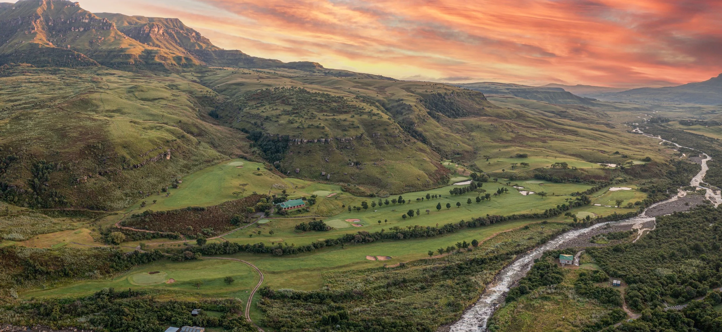A scenic view of Cathedral Peak golf course surrounded by mountains and a river, under a colorful sky at sunset.