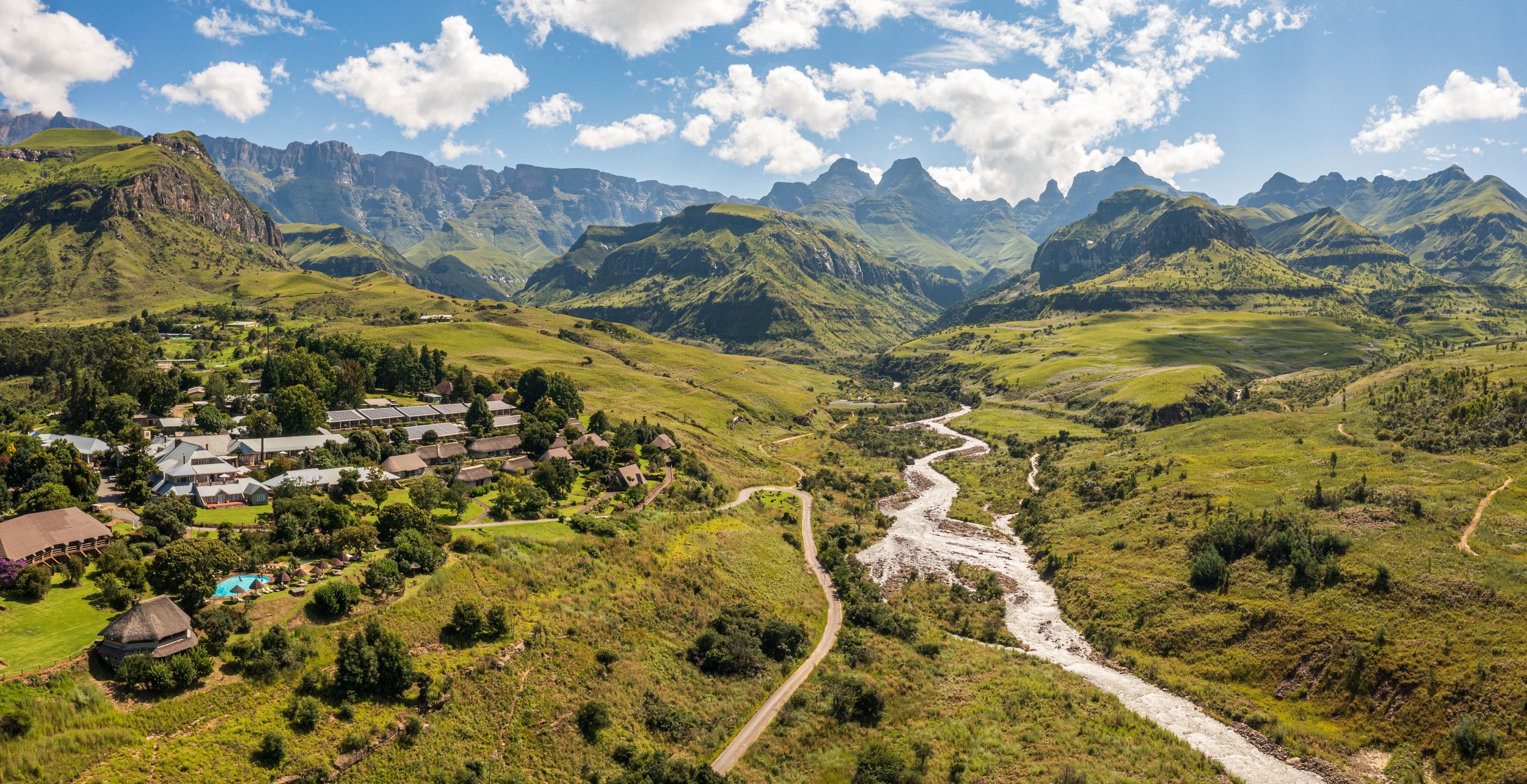 Scenic landscape of a mountain valley with green hills, a river flowing through the center, a small village with houses and trees, and rugged mountains under a partly cloudy sky.