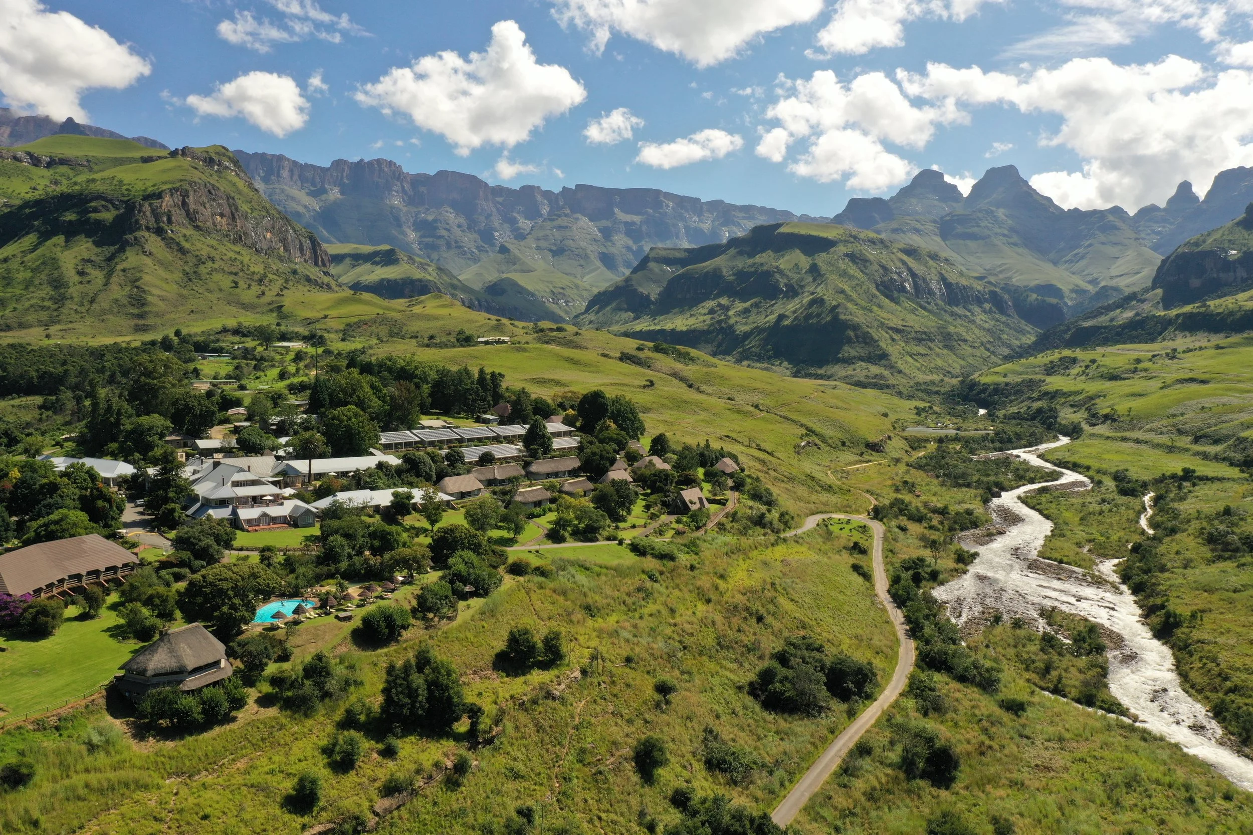 A mountain landscape with lush green hills, a river flowing through the valley, and a small village with buildings, trees, and a swimming pool surrounded by a fence.