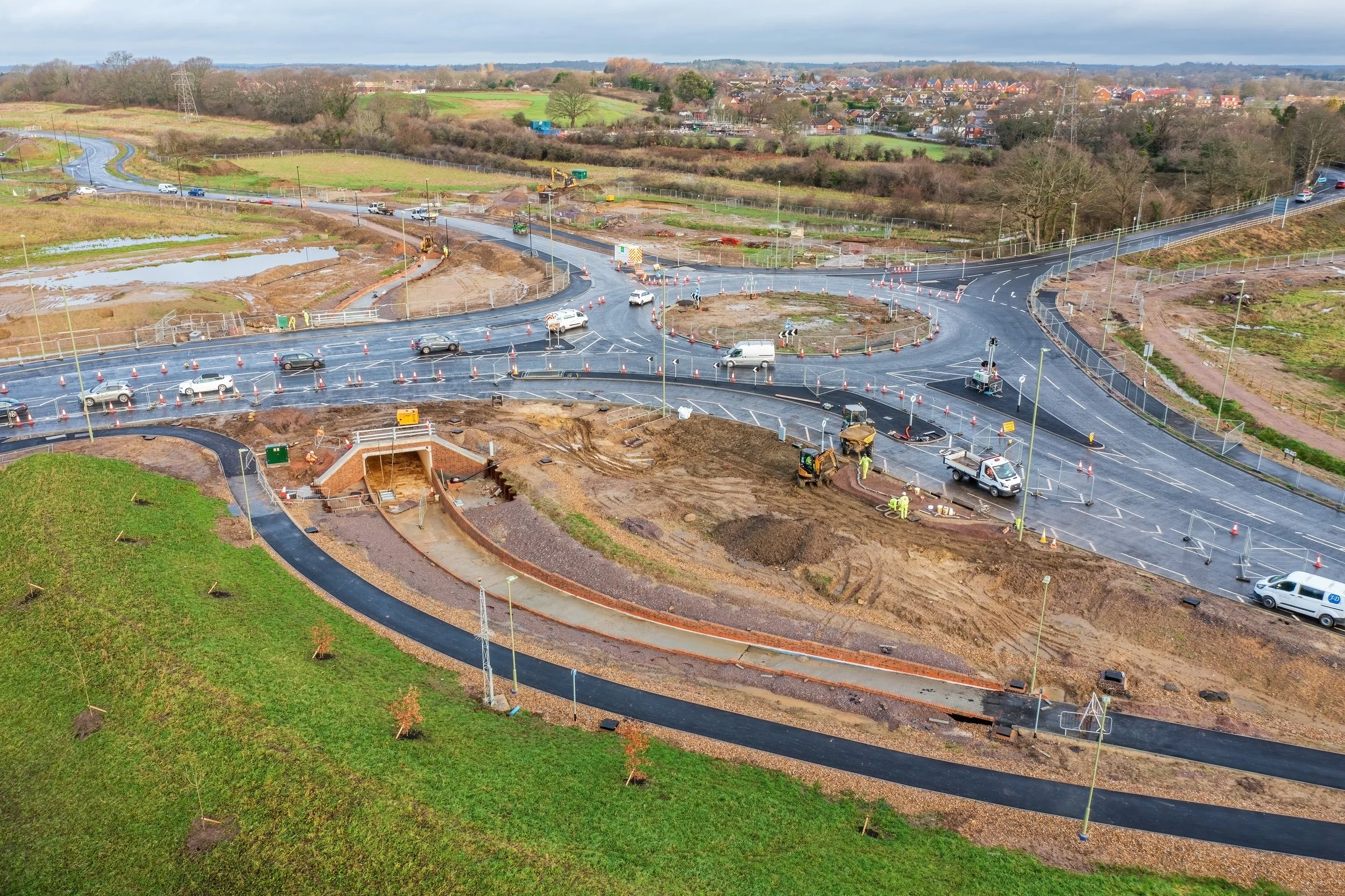 Construction site of a roundabout with multiple lanes and ongoing roadwork, with construction workers and equipment present, adjacent to a grassy area with small trees, and a residential neighborhood in the background under a cloudy sky.