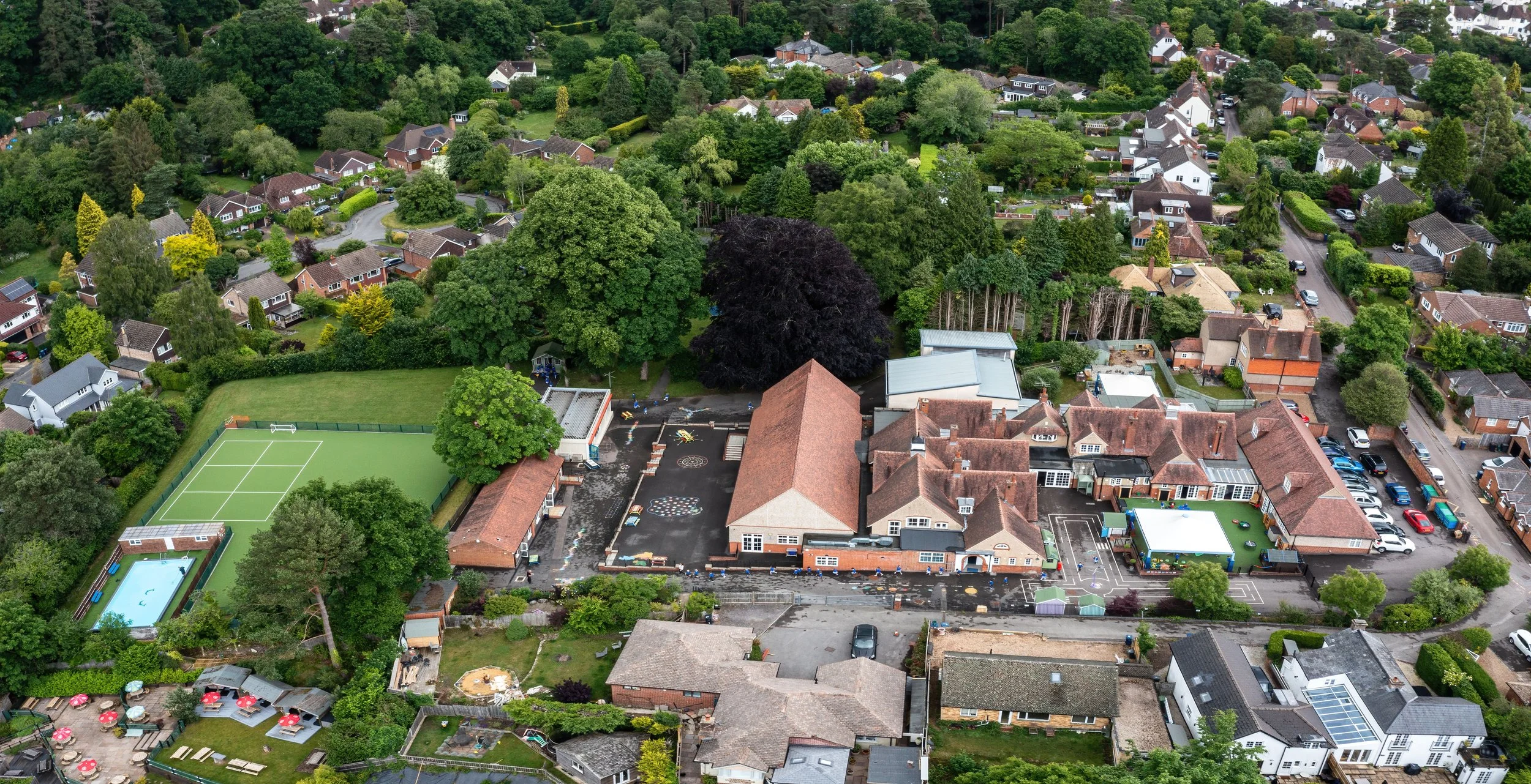 Aerial view of a school campus surrounded by residential houses, trees, and greenery. Features include a tennis court, swimming pool, playground, parking lot, and various buildings.