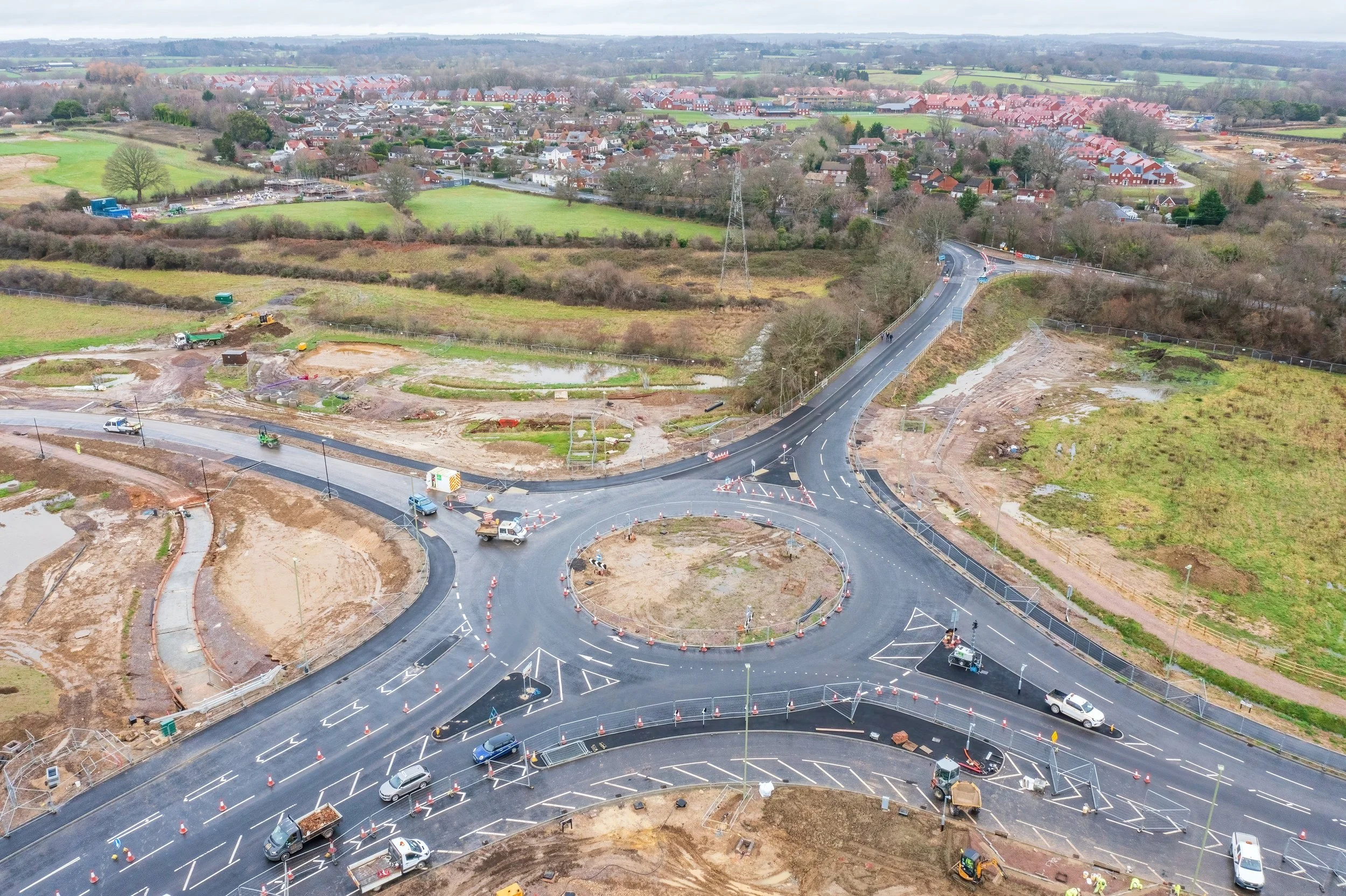 Aerial view of a road construction site with multiple roads, roundabouts, and construction vehicles, surrounded by green fields and a residential area in the background.