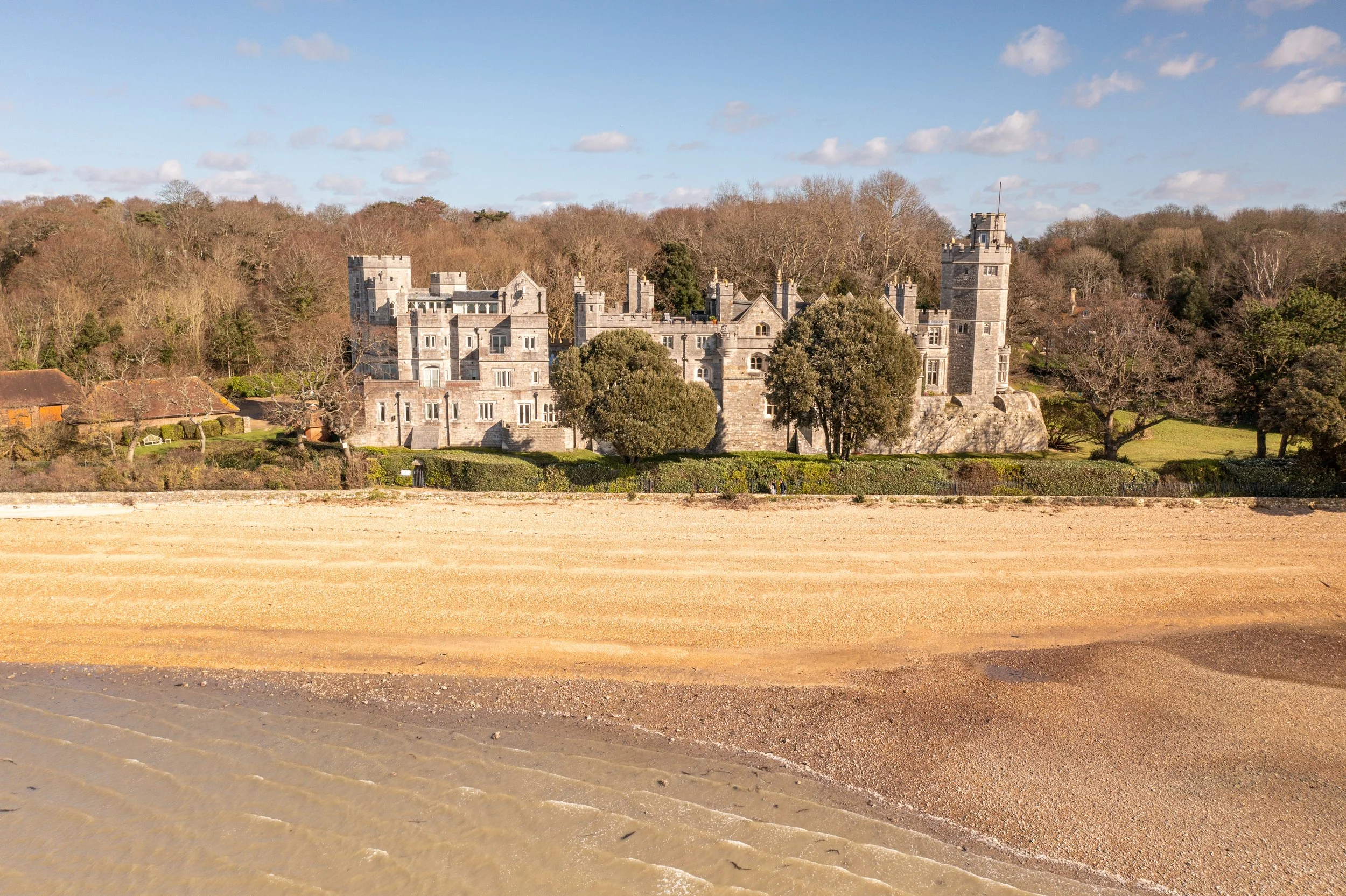 A large stone castle with towers and battlements situated on a hillside, surrounded by trees and greenery, with a beach and water in the foreground under a partly cloudy sky.