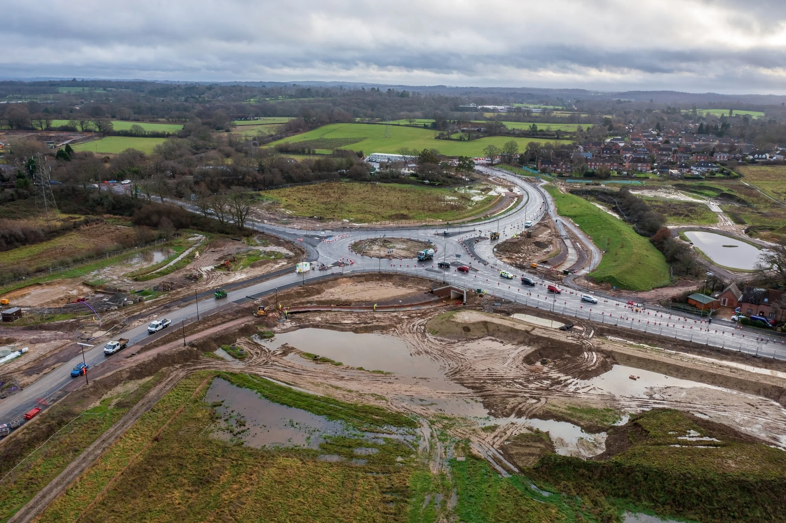 Aerial view of a road construction site with multiple vehicles, dirt, and water-filled areas, surrounded by green fields and a small residential area under an overcast sky.