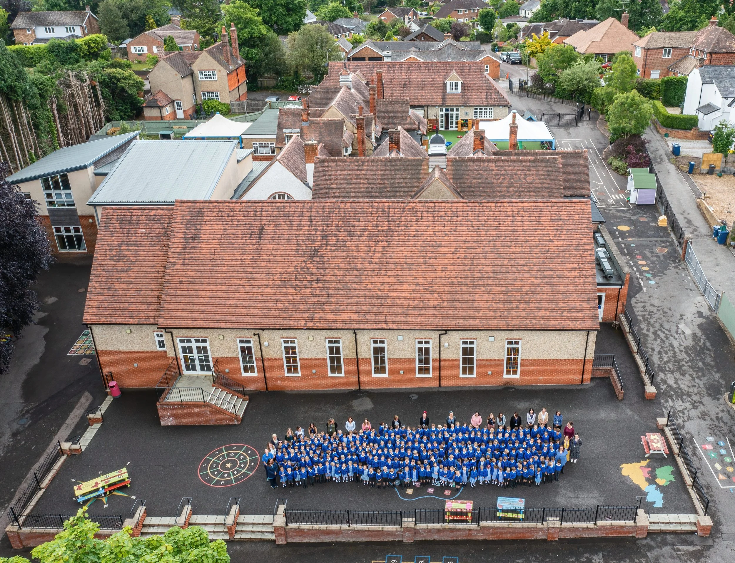An aerial view of a schoolyard with children and adults gathered for a group photo, located in front of a school building with a red-tiled roof, surrounded by residential houses and greenery.