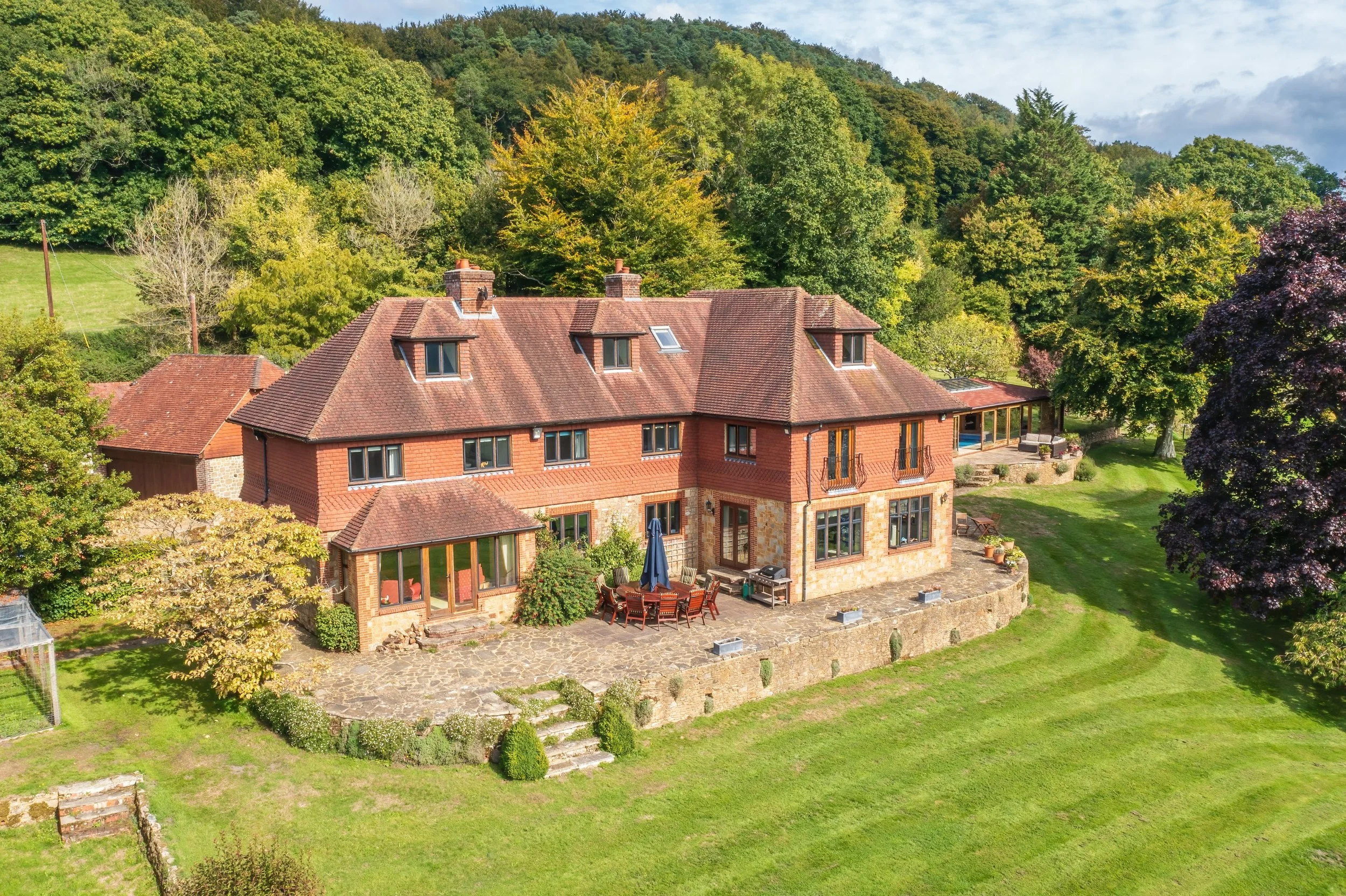 A large brick and stone house with a red-tiled roof, surrounded by lush green trees and lawn, and a dotted patio with outdoor furniture.