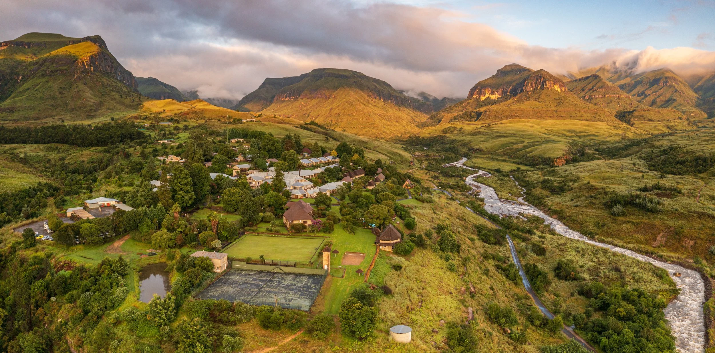 Aerial view of a small village surrounded by lush green mountains and hills, with a winding river running through the landscape, during sunset or sunrise.